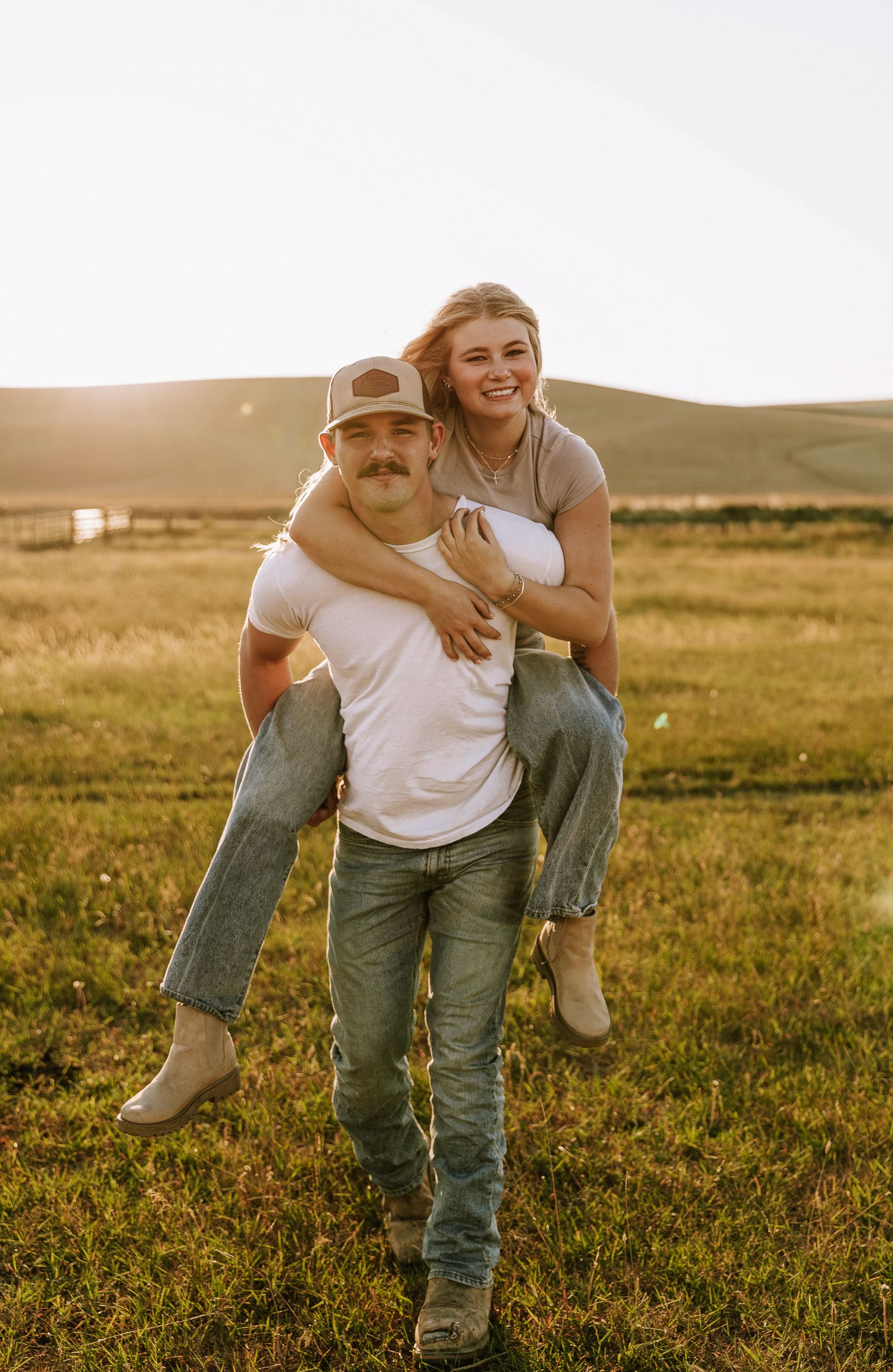 A couple shares a playful piggyback ride moment in a golden field at sunset.