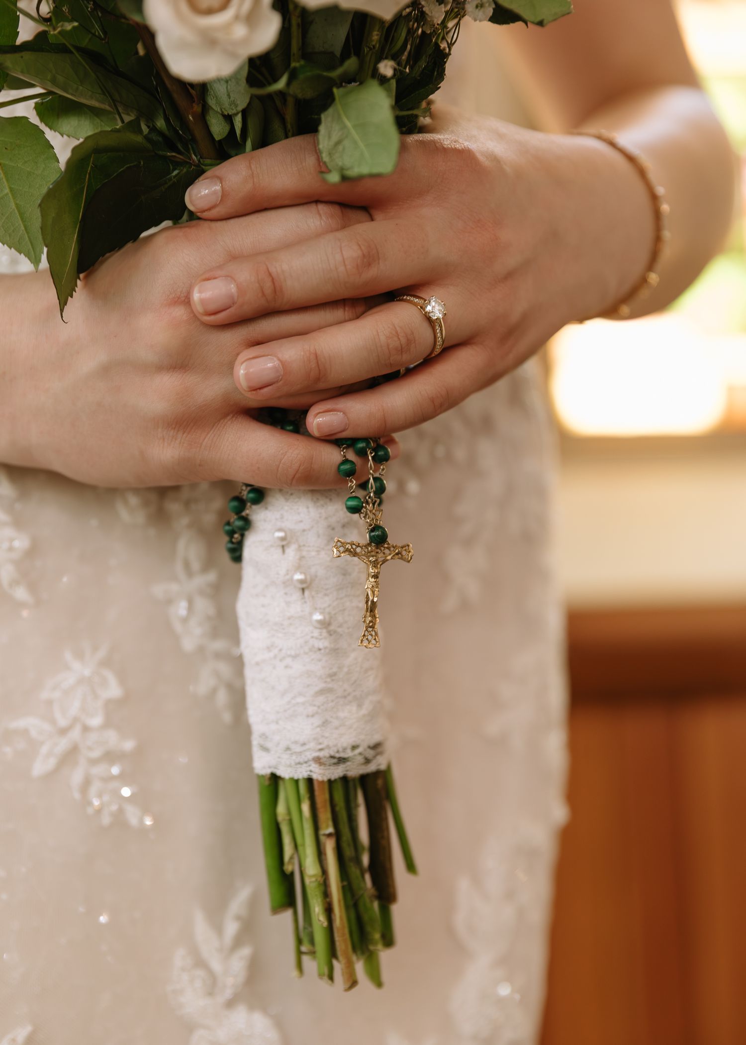 Close up of hands holding white rose bouquet wrapped in lace, showing engagement ring and rosary detail.
