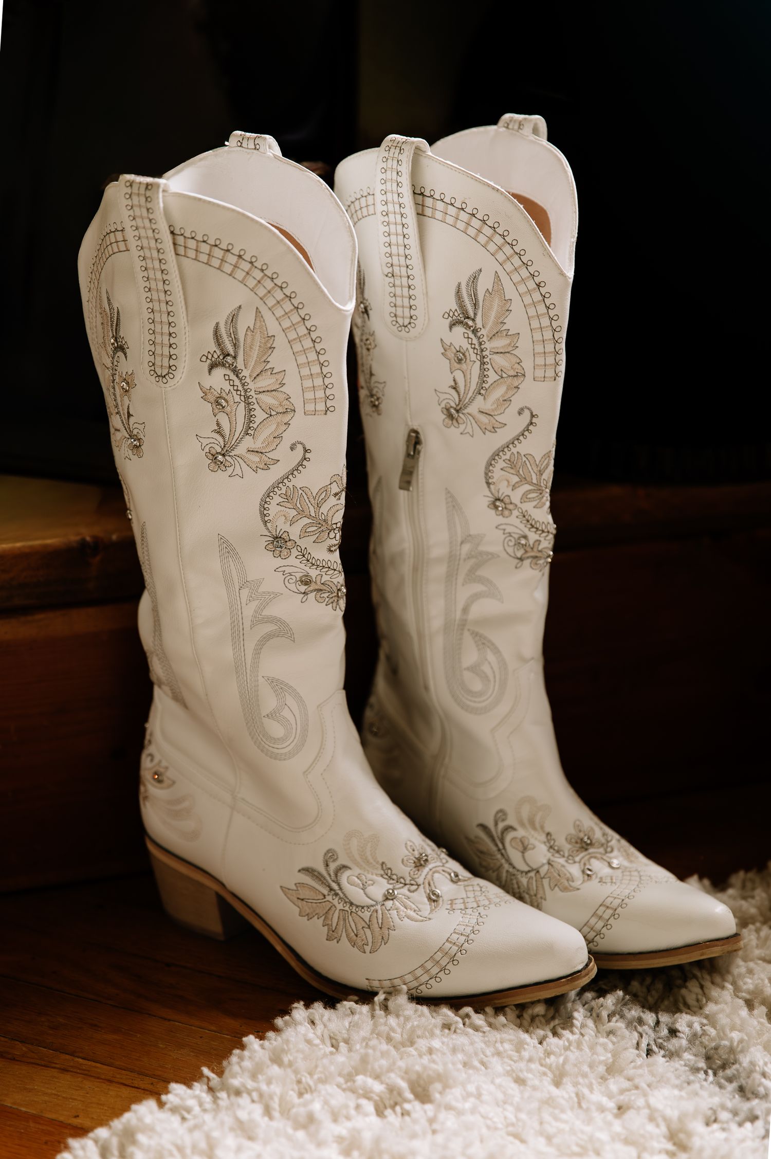 White leather cowboy boots with intricate silver embroidery designs displayed on a dark wooden floor with white decor.