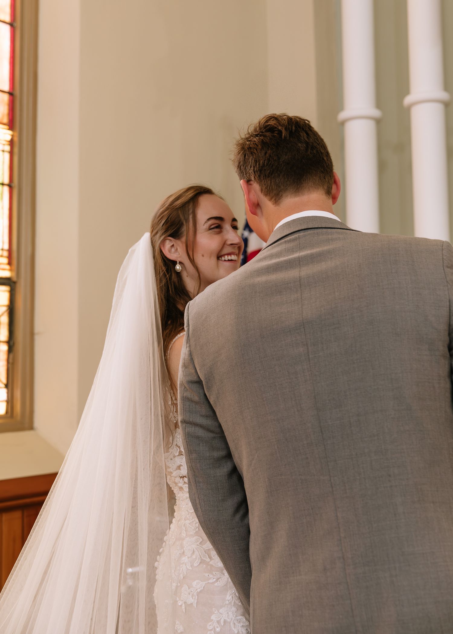 Bride and groom share a candid laughing moment in an elegant church setting with white pillars and natural lighting.