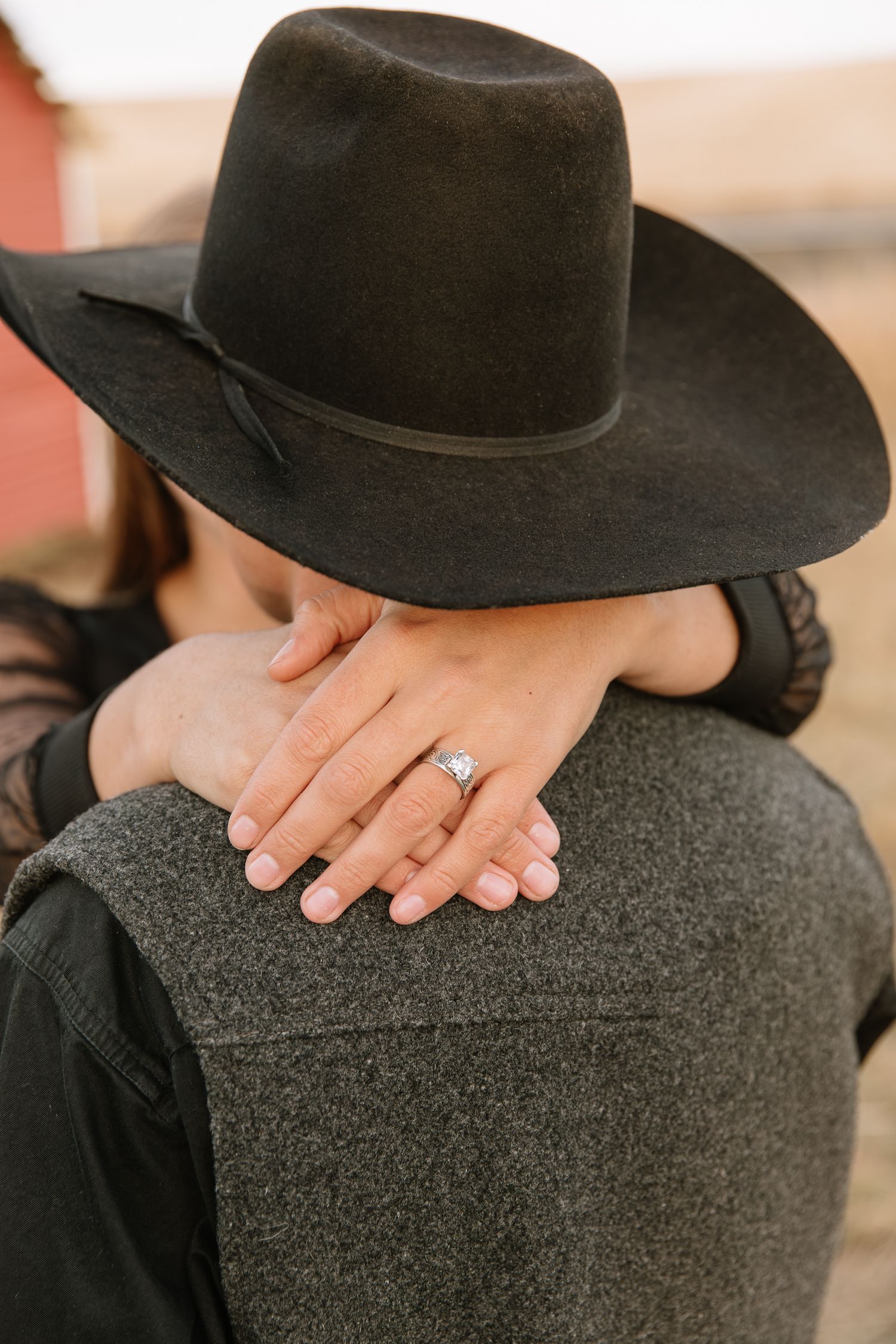 A hand wearing an engagement ring rests on a gray wool surface next to a black wide-brimmed hat.