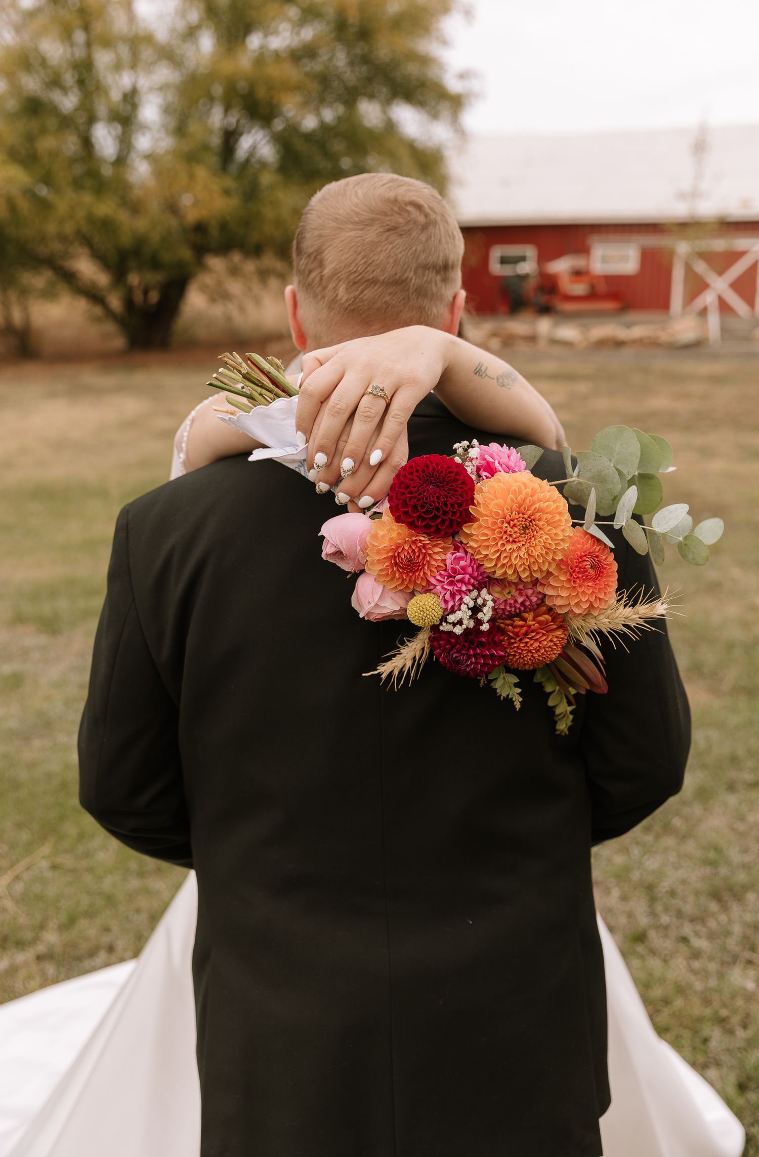 A couple embraces from behind with vibrant orange and pink floral arrangements decorating their shoulders in an outdoor setting.