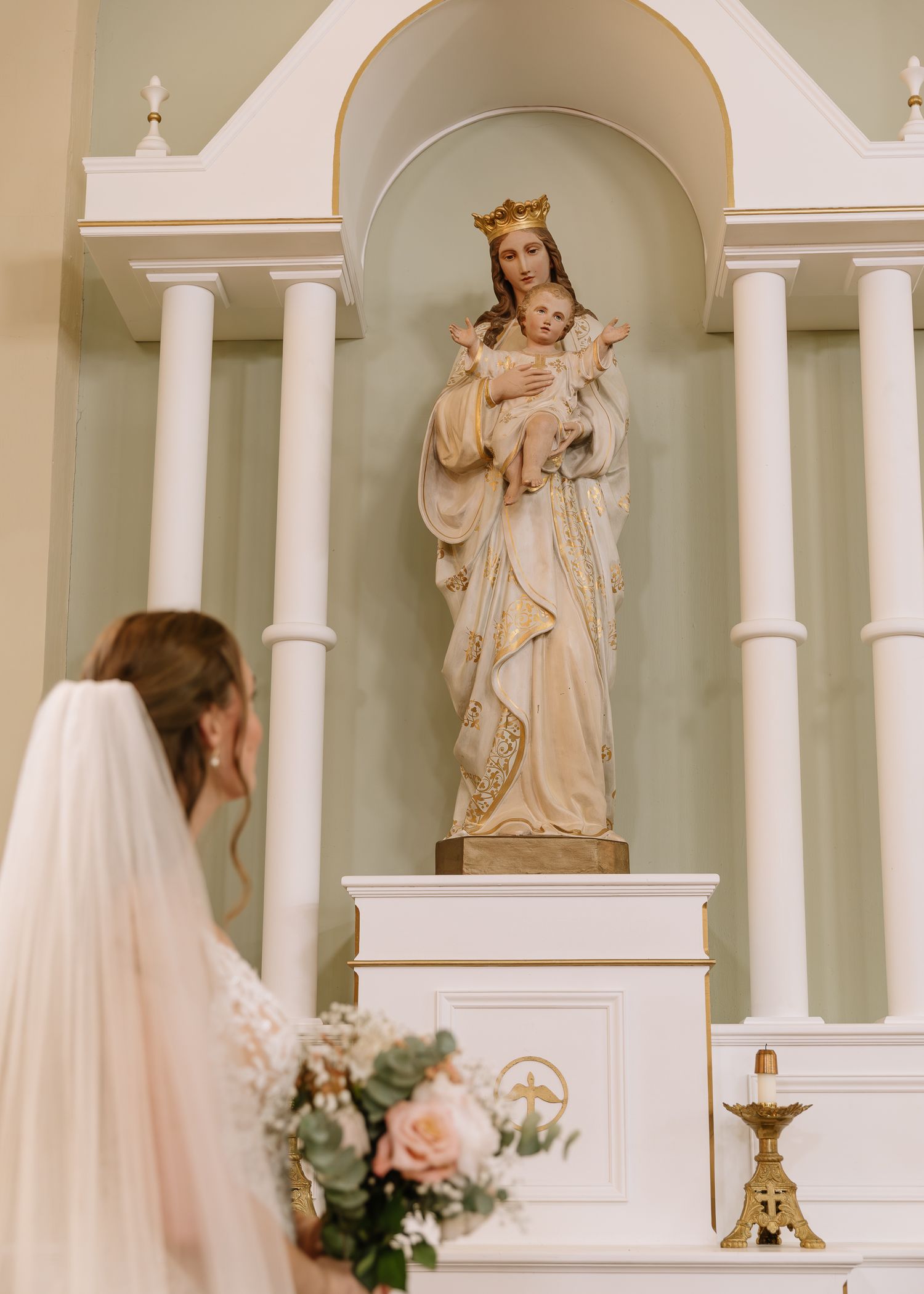 A wedding ceremony takes place in an elegant white church interior with arched columns and religious statues.