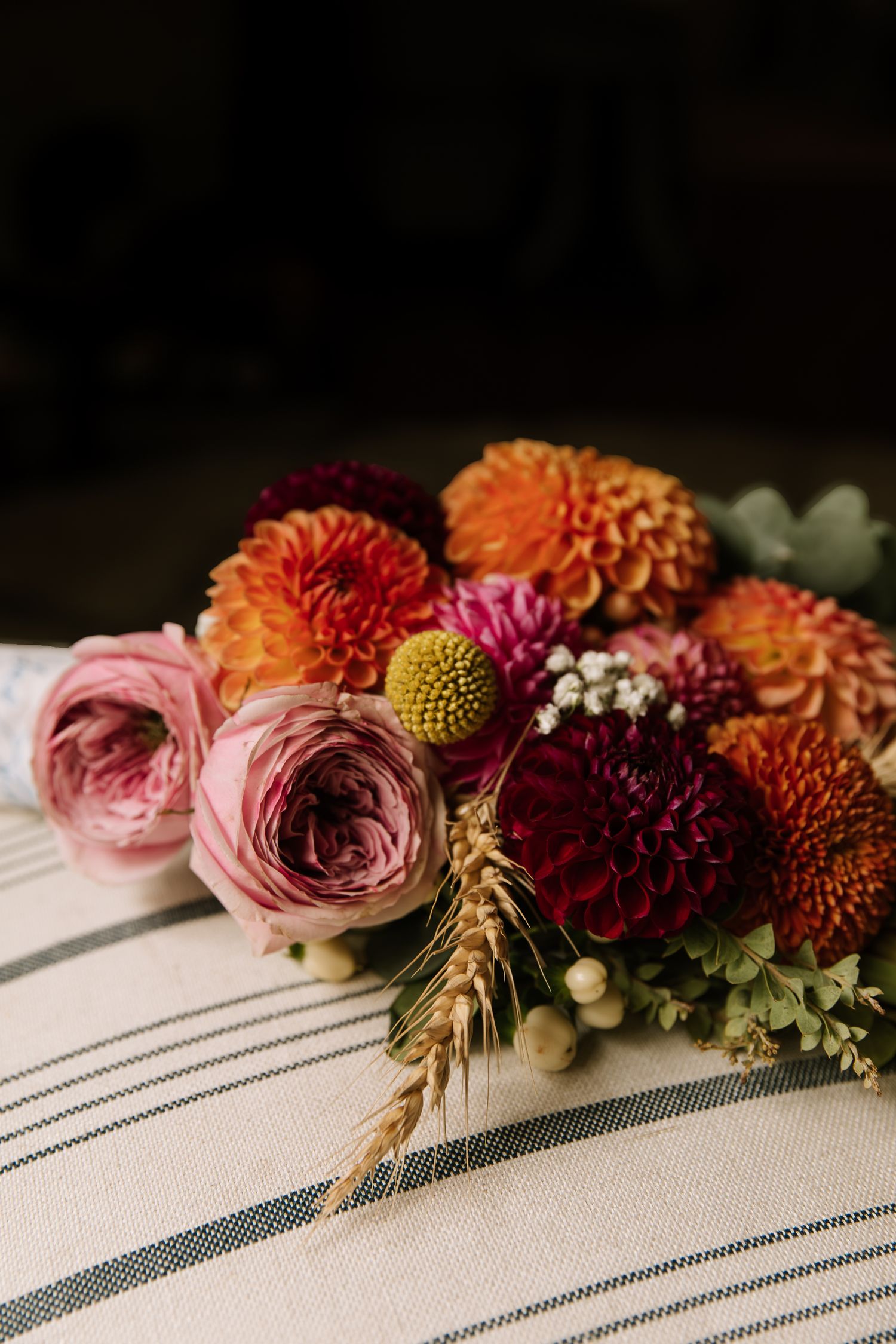 Beautiful fall floral arrangement with pink ranunculus, orange dahlias, and wheat stalks on striped linen fabric.