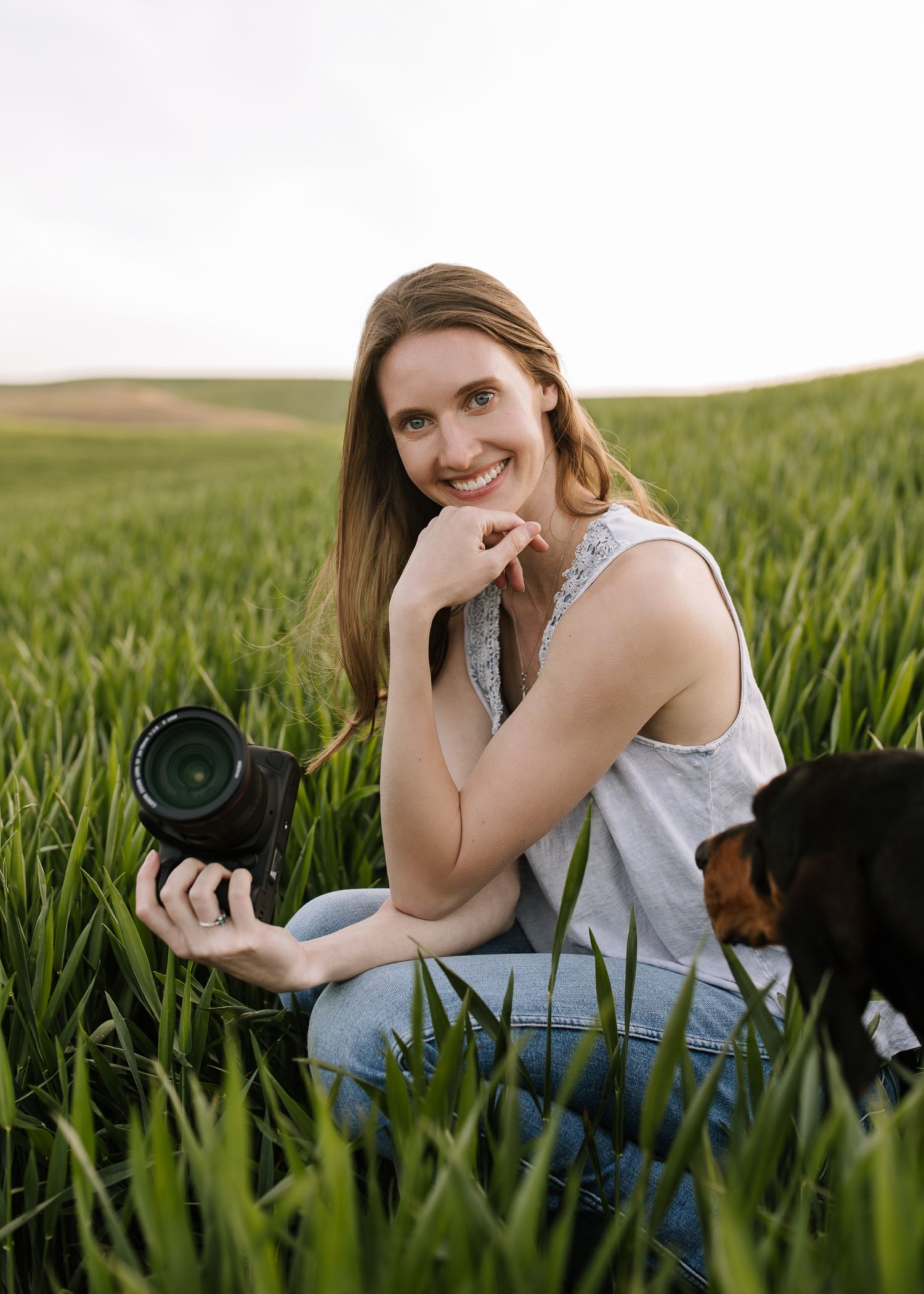 A photographer in a white top lies in a lush green field while holding a professional camera.