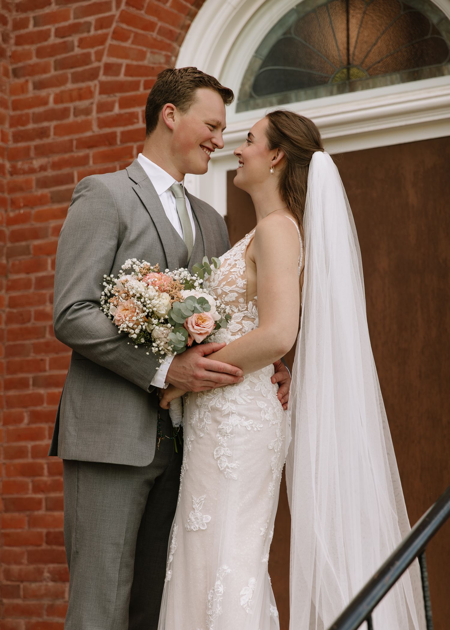 A bride and groom share intimate moments outside a brick church building while holding a peach and white floral bouquet.