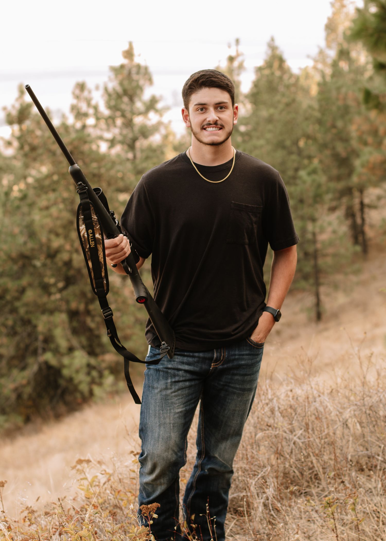 Person in black t-shirt and jeans standing in grassy field holding rifle in various poses.