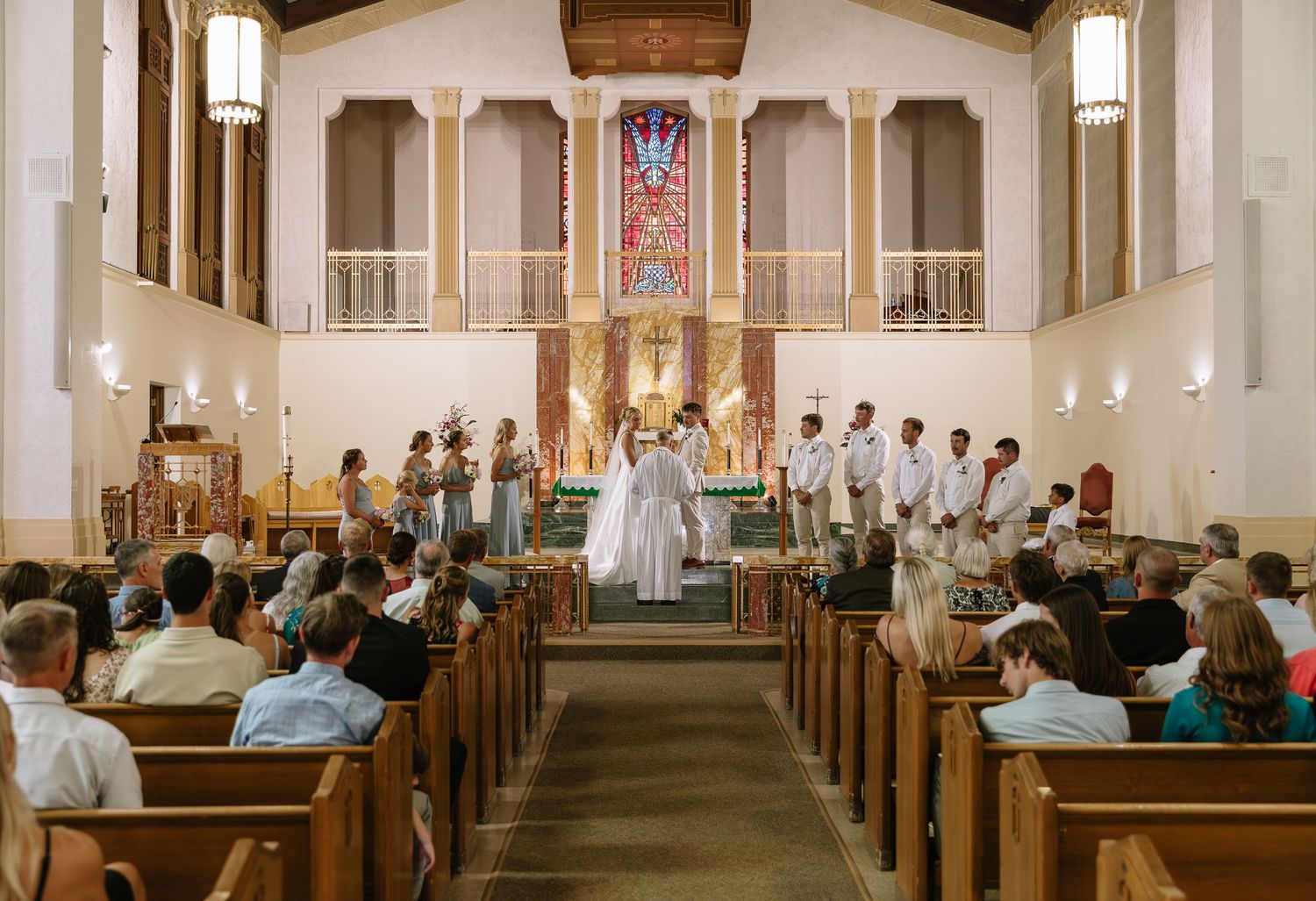 People sit in wooden pews inside a traditional church sanctuary with exposed wooden beams and stained glass windows.
