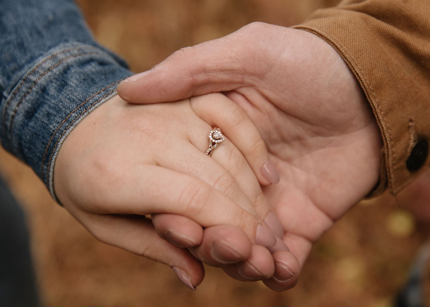 Close-up of two hands clasped together with an engagement ring, wearing denim and brown jacket sleeves.