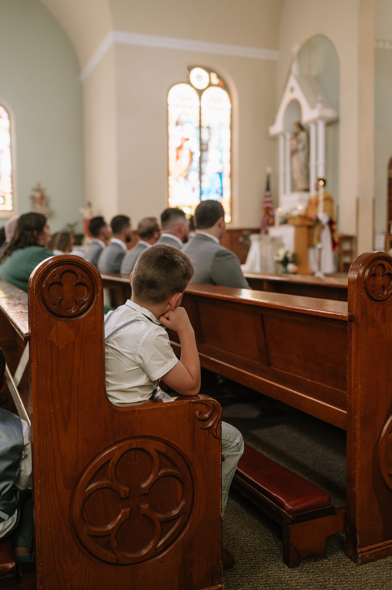 Young students in school uniforms sit in ornate wooden pews during a church service with stained glass windows visible.