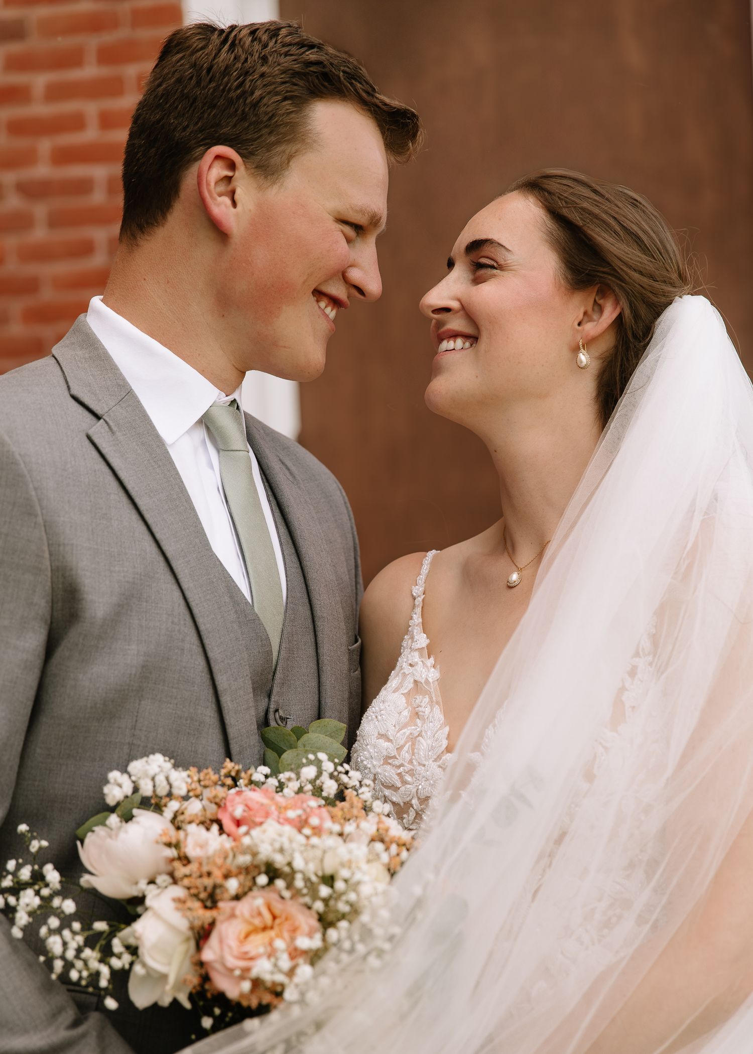 A bride and groom share joyful moments against a brick wall backdrop while holding a peach and white floral bouquet.