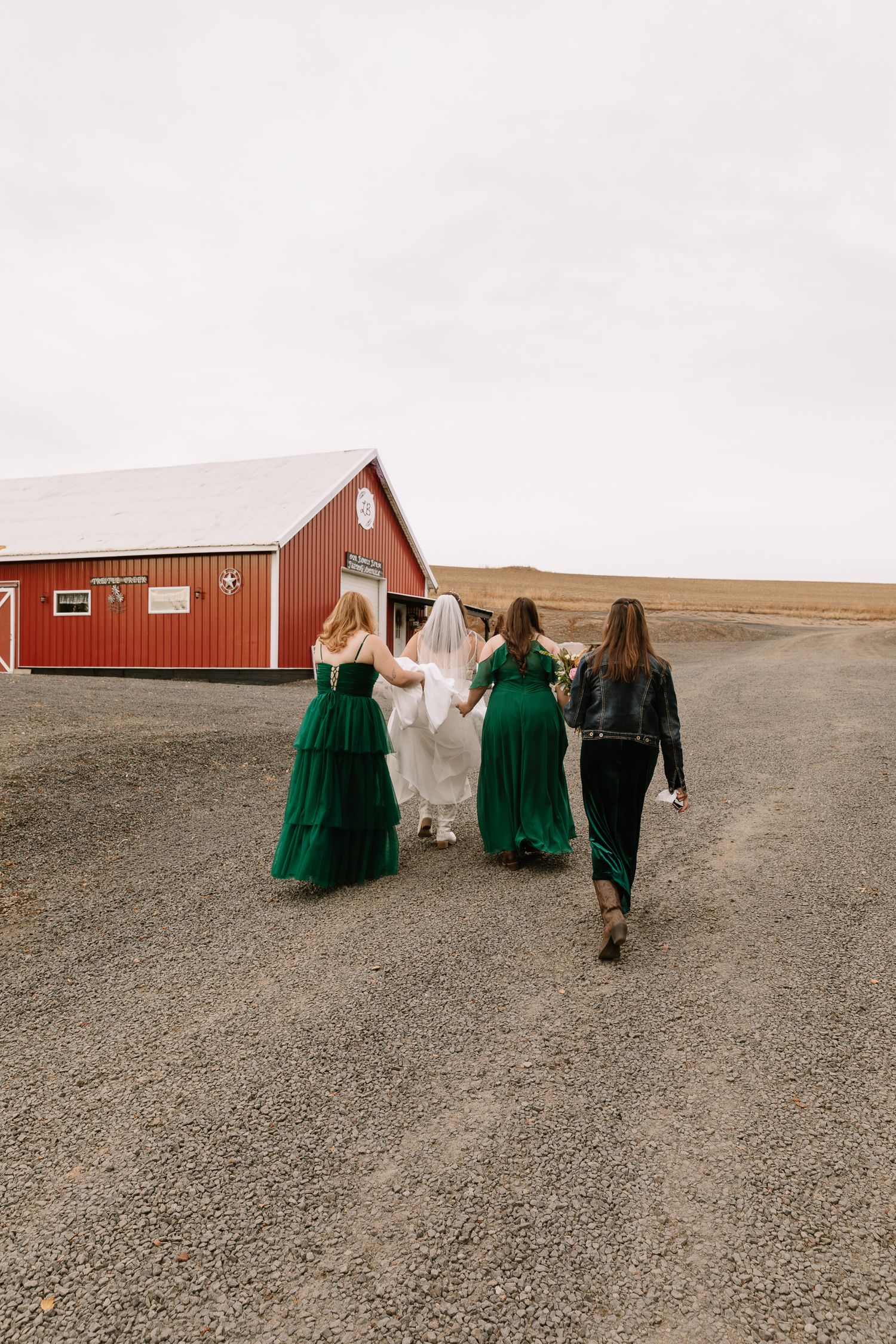 A wedding party in emerald green and white dresses walks along a gravel path toward red barns in a rural setting.