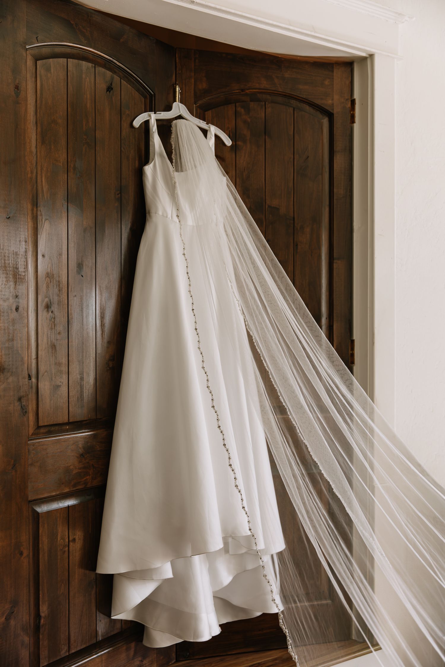 A white wedding dress and veil hanging against dark wooden panels in natural light.