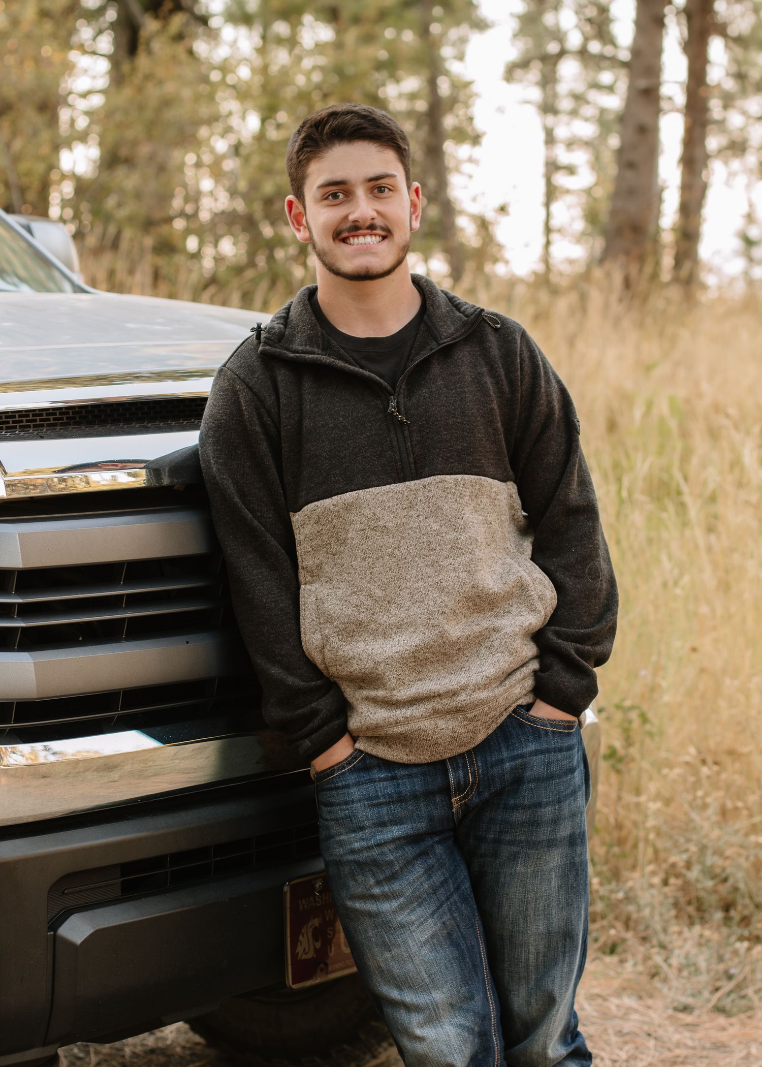 A person wearing a black hoodie and jeans poses in front of a truck in a wooded outdoor setting.