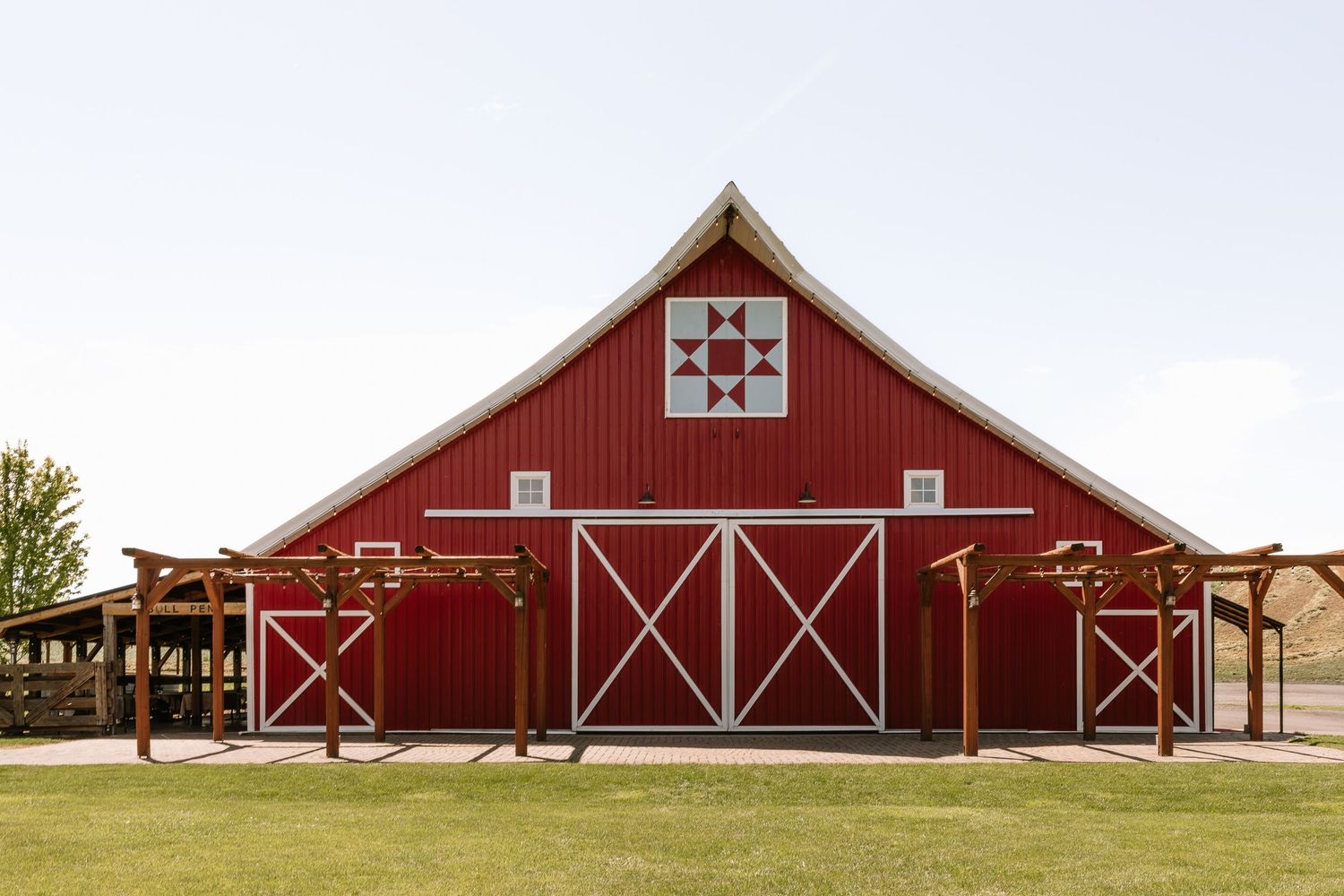 A red barn with white trim and X-shaped doors stands against a clear blue sky in a rural landscape.
