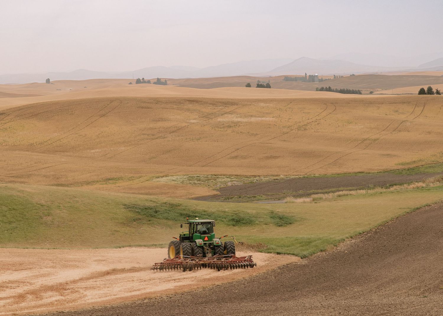 Green tractor plowing through rolling wheat fields in the Palouse region of eastern Washington on a hazy day.