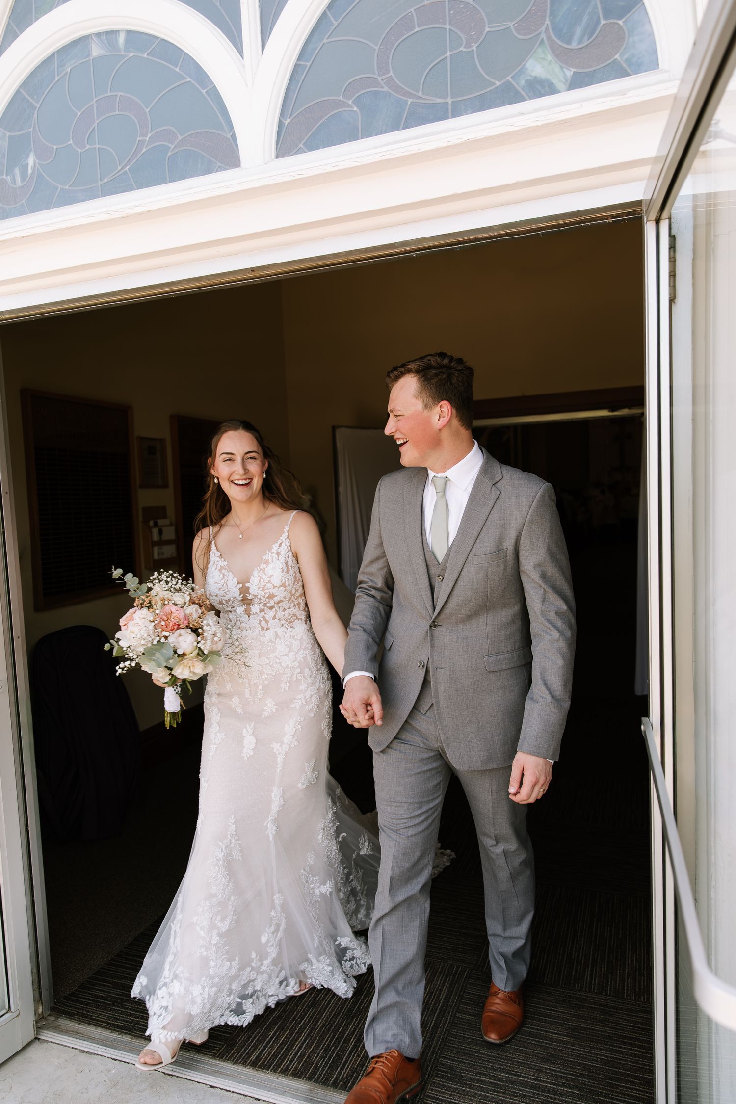 A bride and groom walk together through an arched hallway with white architectural details and natural lighting.
