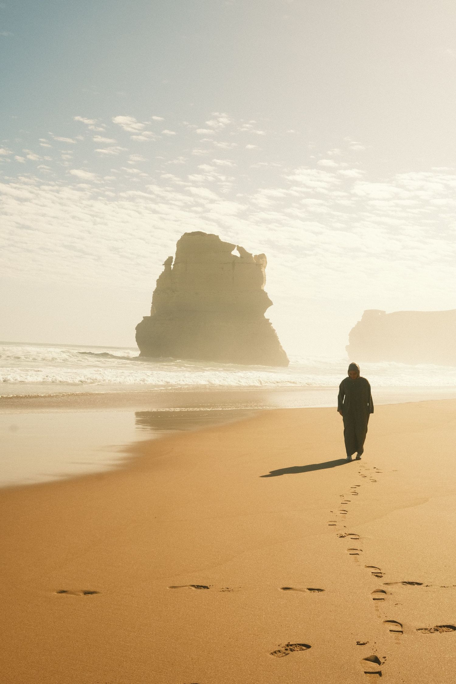 Silhouette of a person walking along a golden beach with large rock formations in the distance.