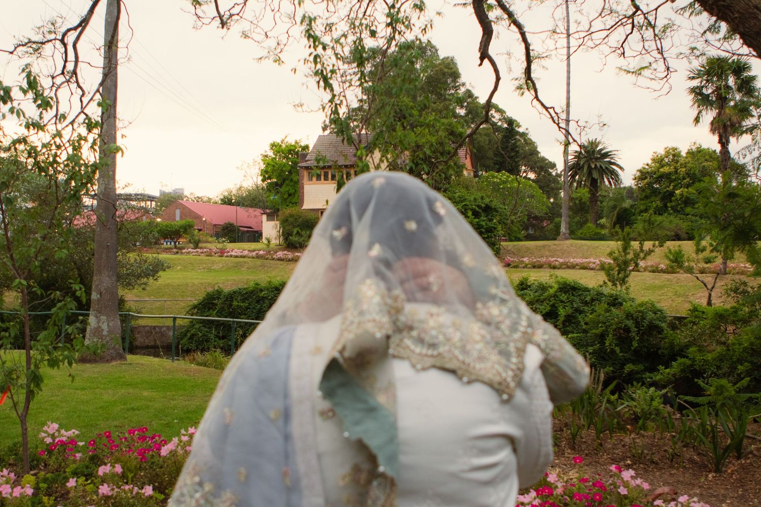 A bride in a white dress and veil stands in a garden setting during sunset with flowers and trees in the background.