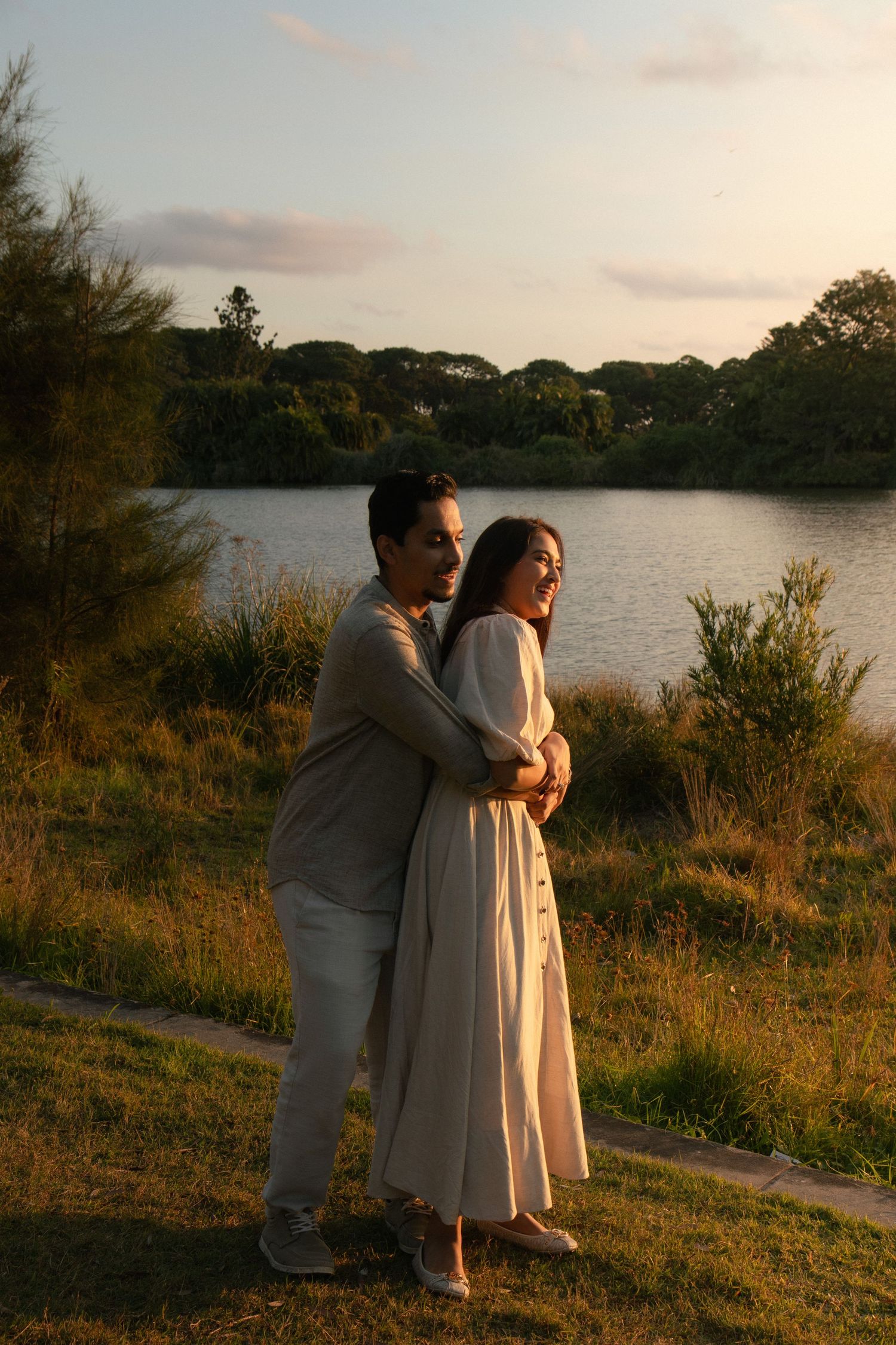Couple embracing at golden hour by a serene lake, surrounded by lush greenery, woman in white dress, romantic outdoor portrait.