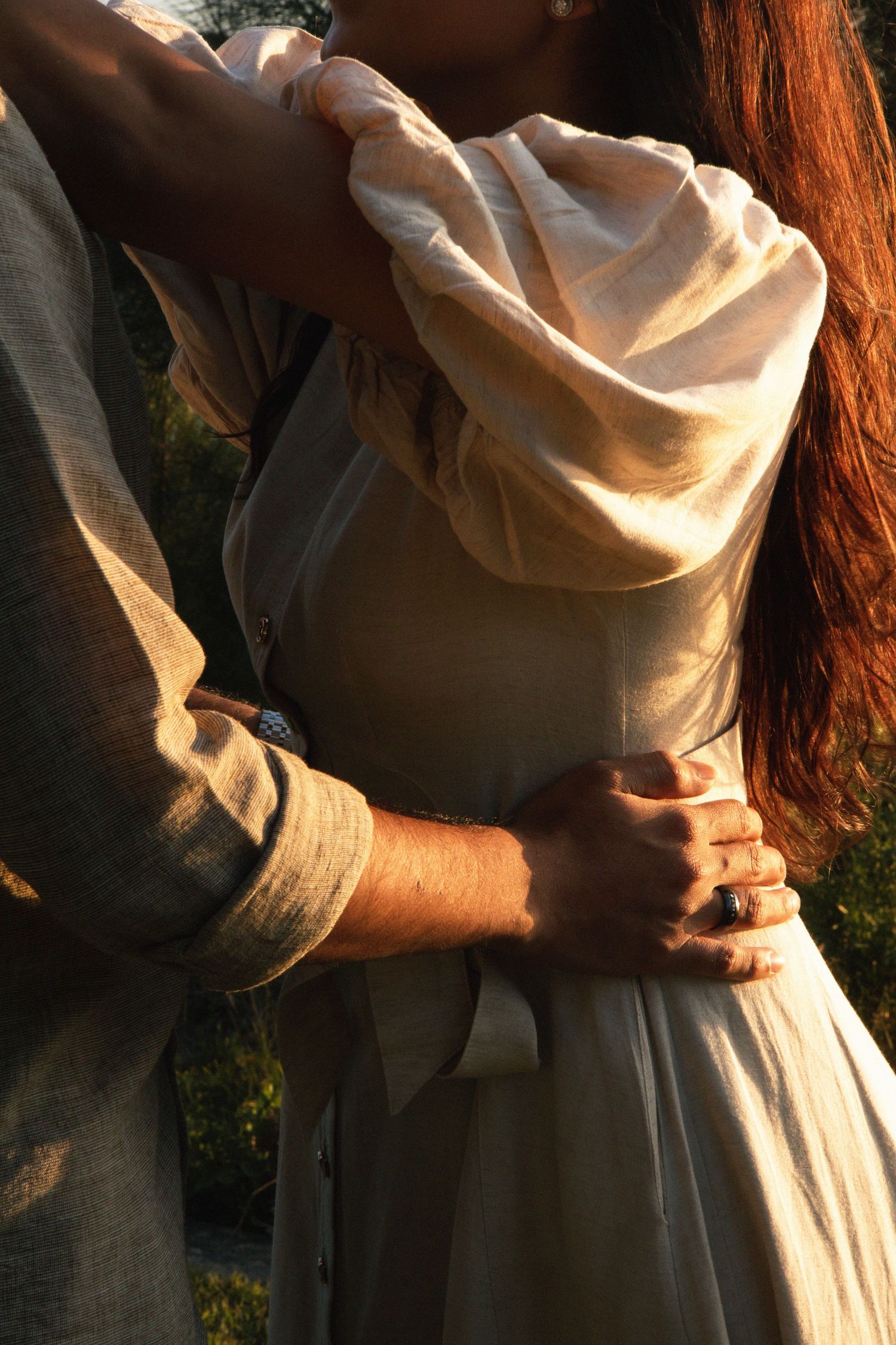Couple embracing outdoors in warm golden light, wearing linen clothing, man's arm around woman's waist.