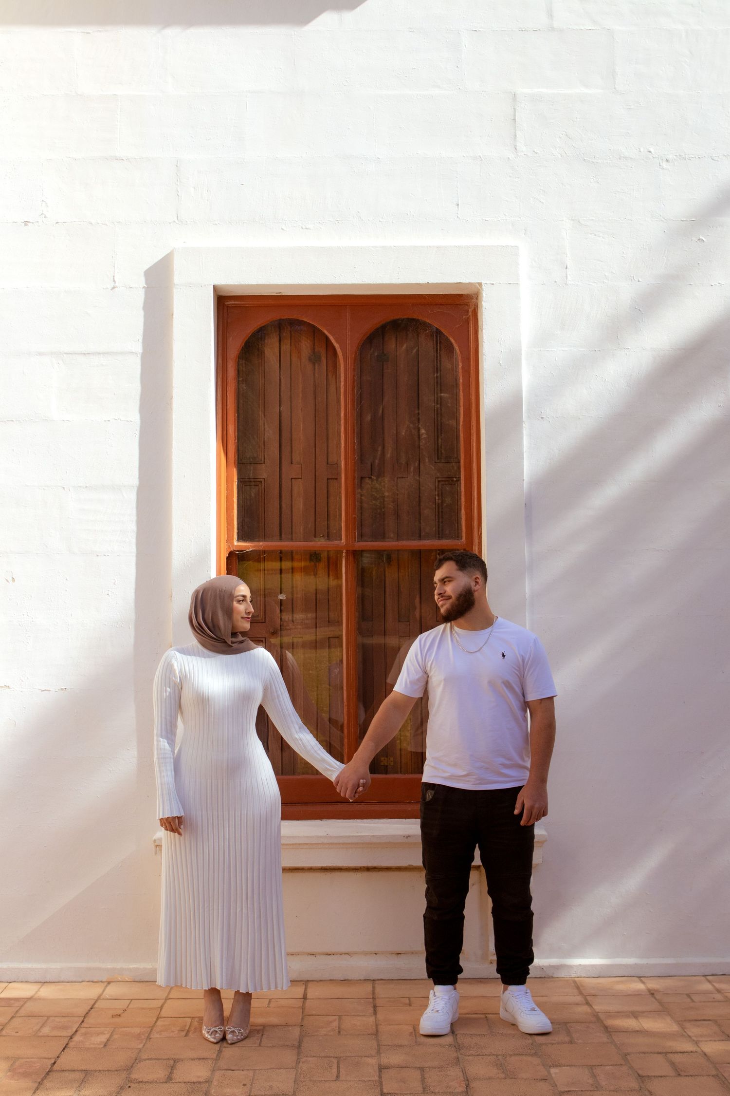A couple wearing white and black stands on a tiled terrace with wooden windows.