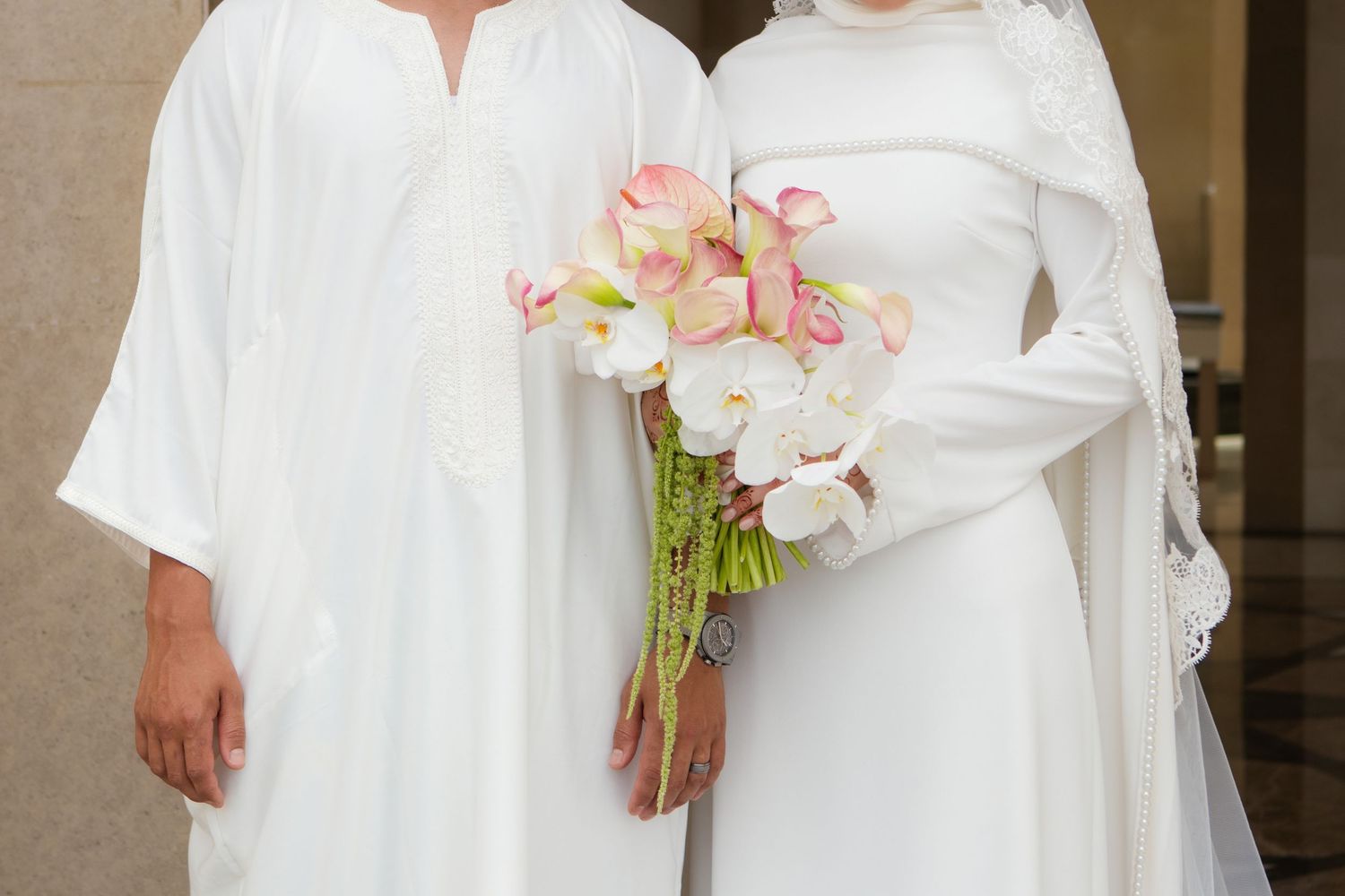 Two people in white attire pose together, one holding a bouquet of pink and white flowers at a wedding.
