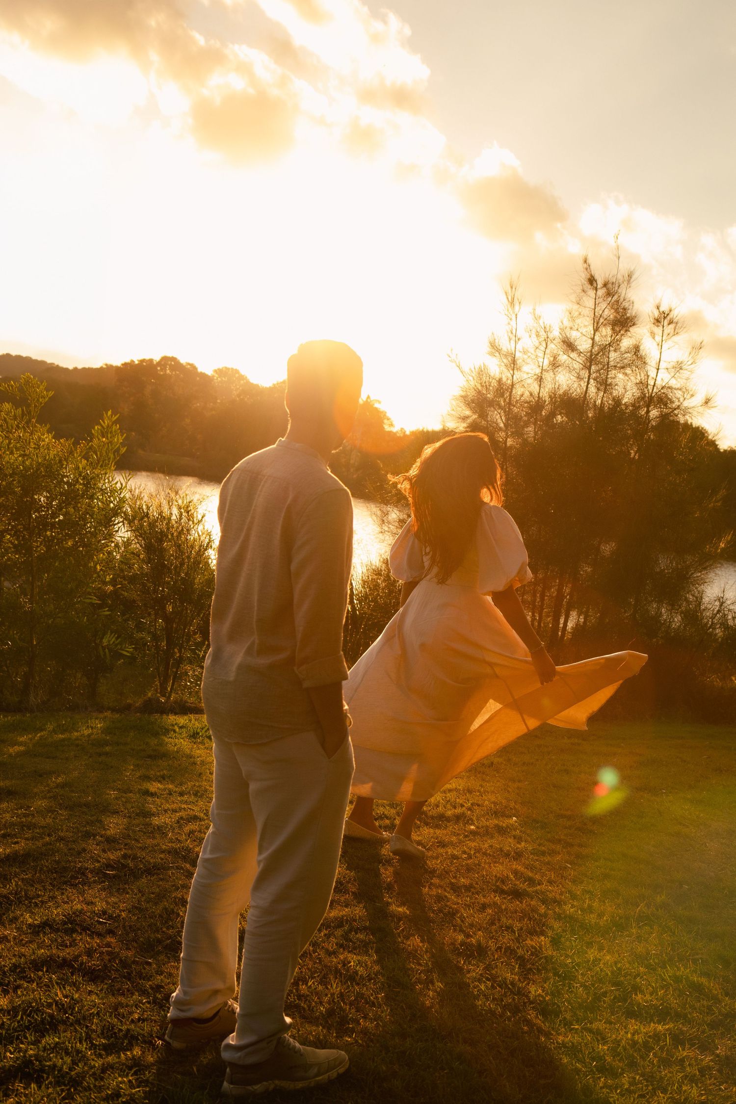 Couple standing together at golden sunset by a lake, woman in white dress with light flare, romantic outdoor scene.