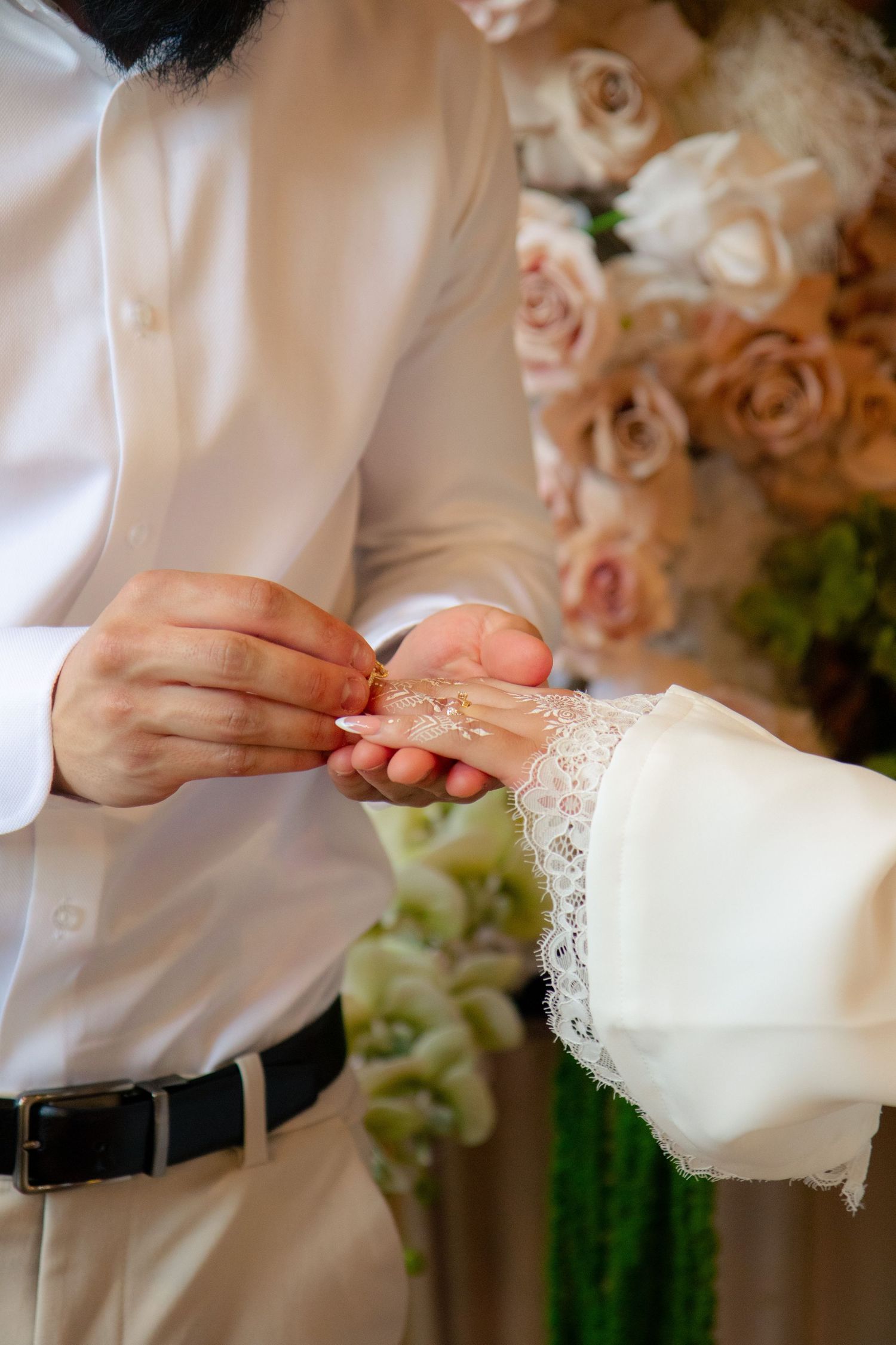 Groom placing a wedding ring on bride's lace-sleeved hand at floral ceremony backdrop.