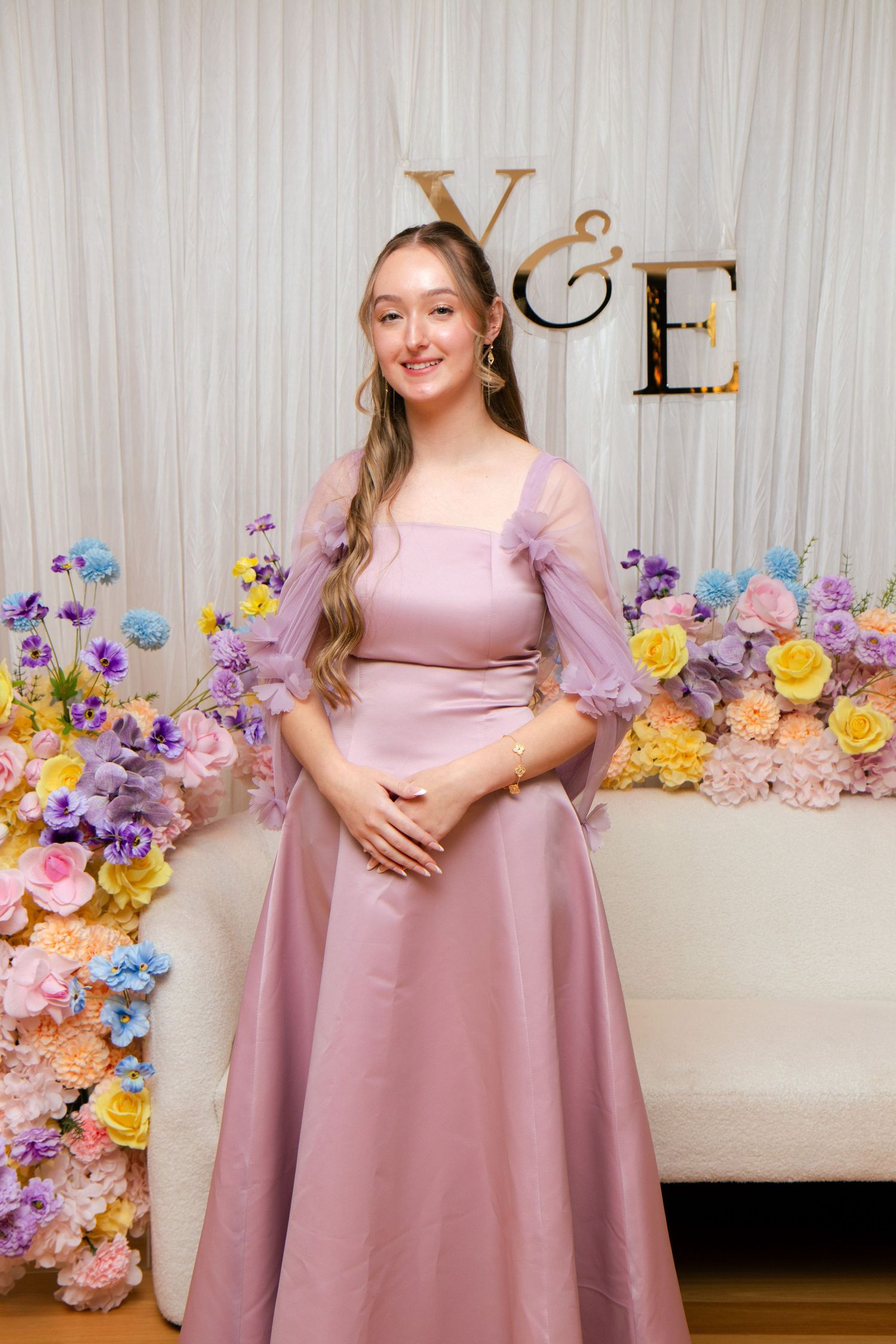 A young woman in a mauve satin gown with sheer sleeves poses before a floral backdrop at a Y&E event.