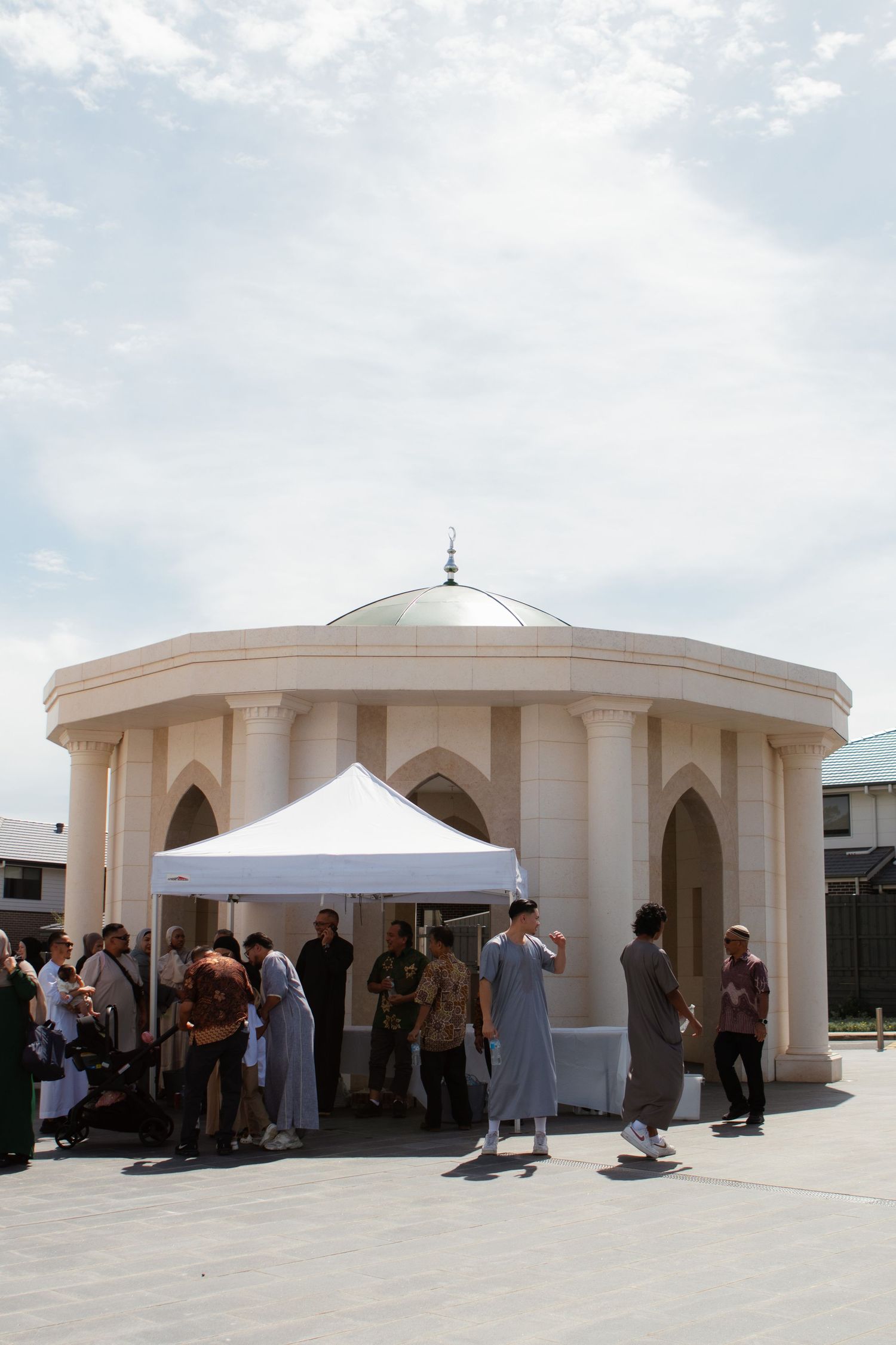 People gather under a white tent next to a domed circular building with arched architectural features on a sunny day.