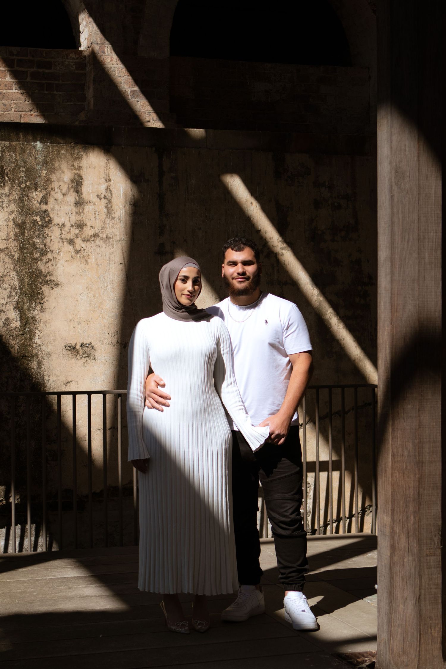 A couple in white and black outfits poses against a dark architectural backdrop with dramatic shadows.