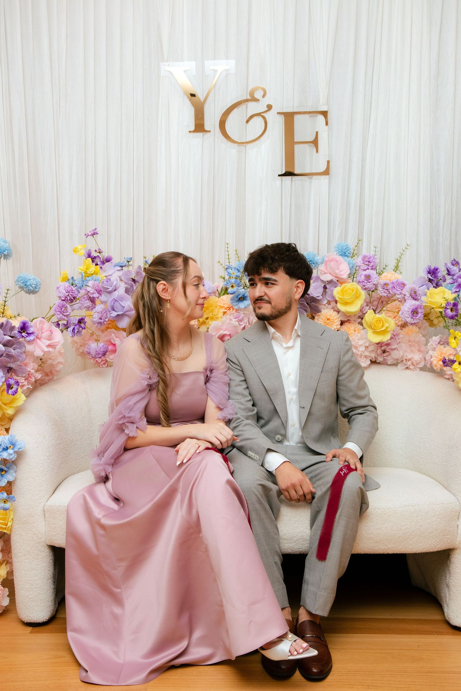Couple in formal attire sitting on a white sofa with colorful floral backdrop and Y&E monogram sign.