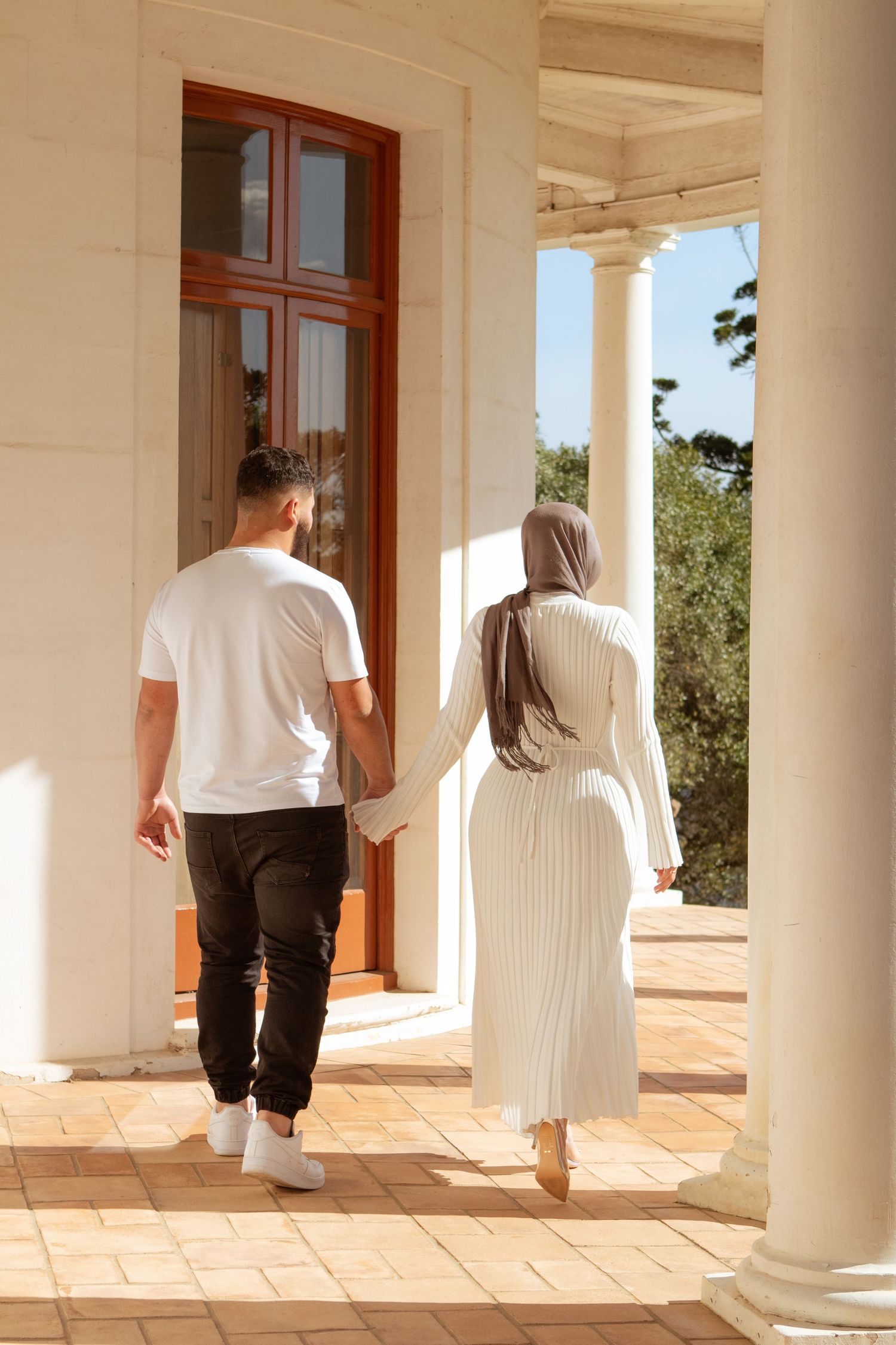 Two people walking along a colonnade with terra cotta flooring and white columns.