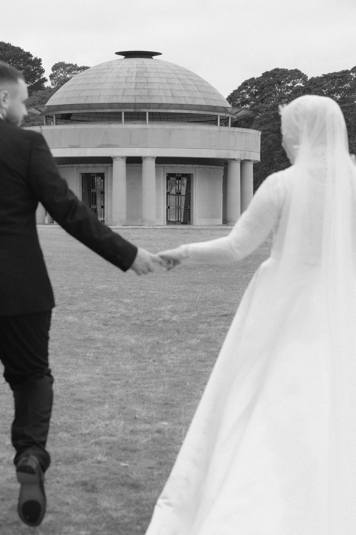 Black and white photo of a bride and groom holding hands walking toward a domed rotunda building on their wedding day.