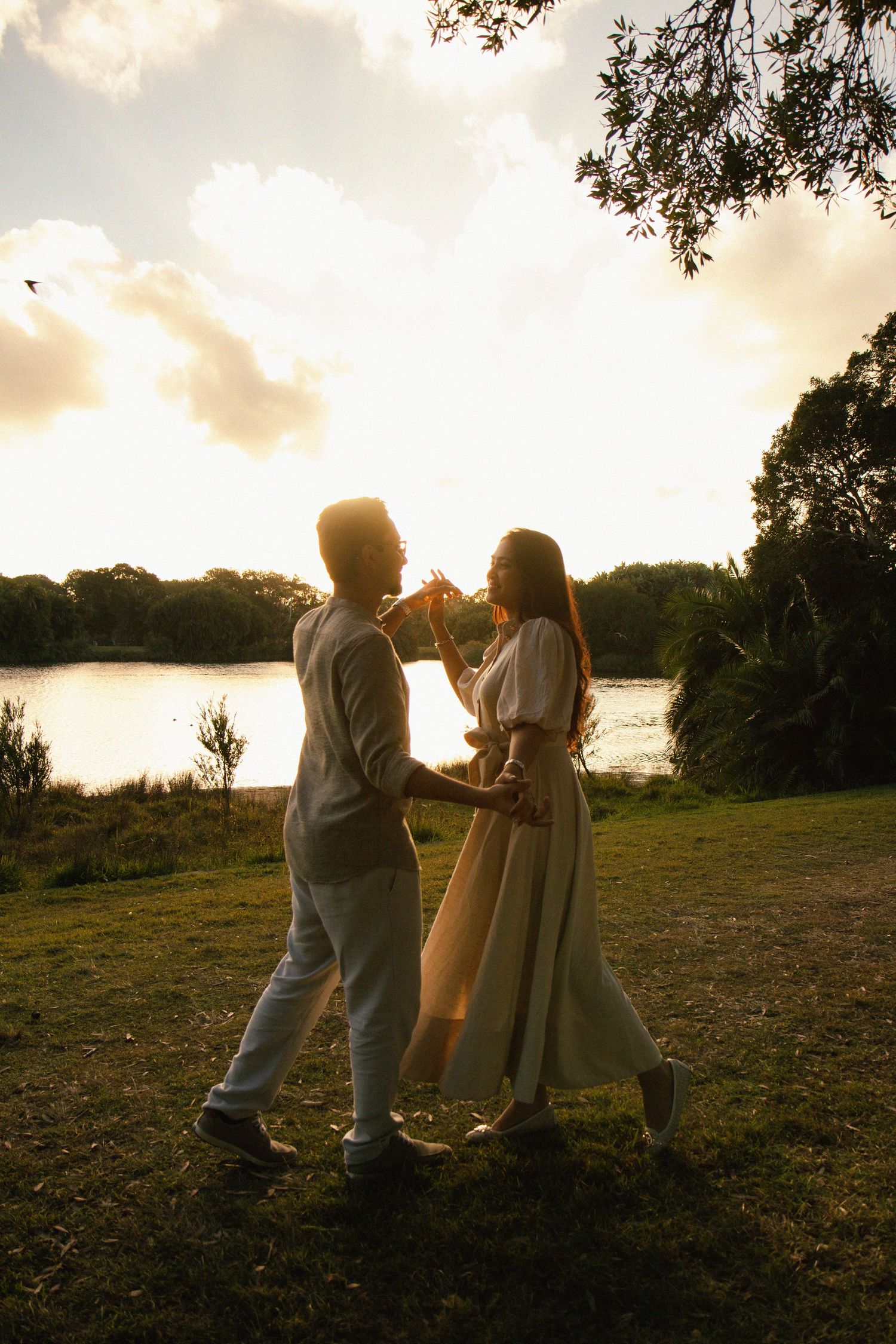 Couple dancing by a sunlit lake at golden hour, surrounded by lush greenery in a romantic outdoor setting.