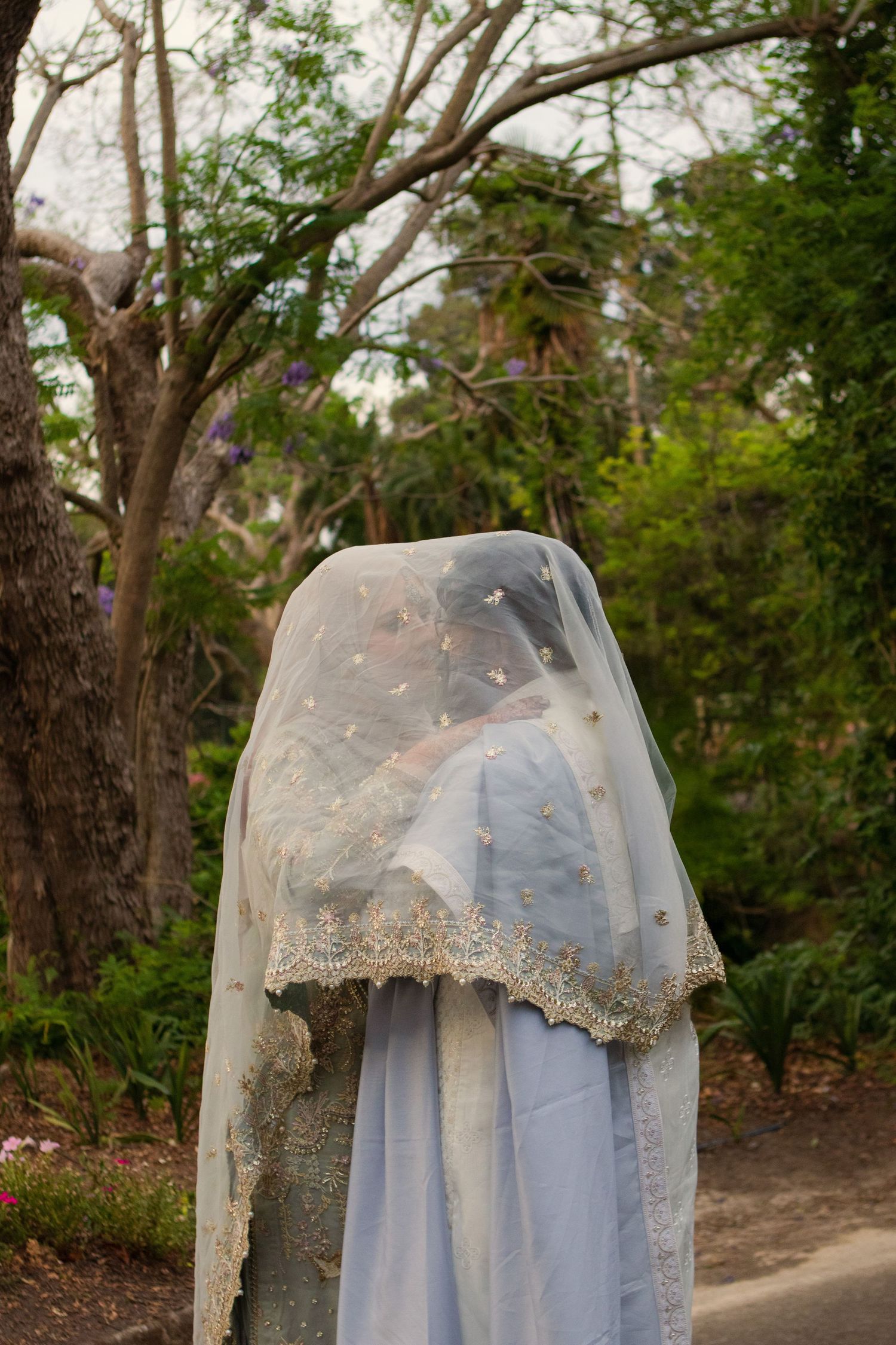 Bride in elegant gray lehenga with gold trim, draped in sheer veil, standing in lush garden setting.
