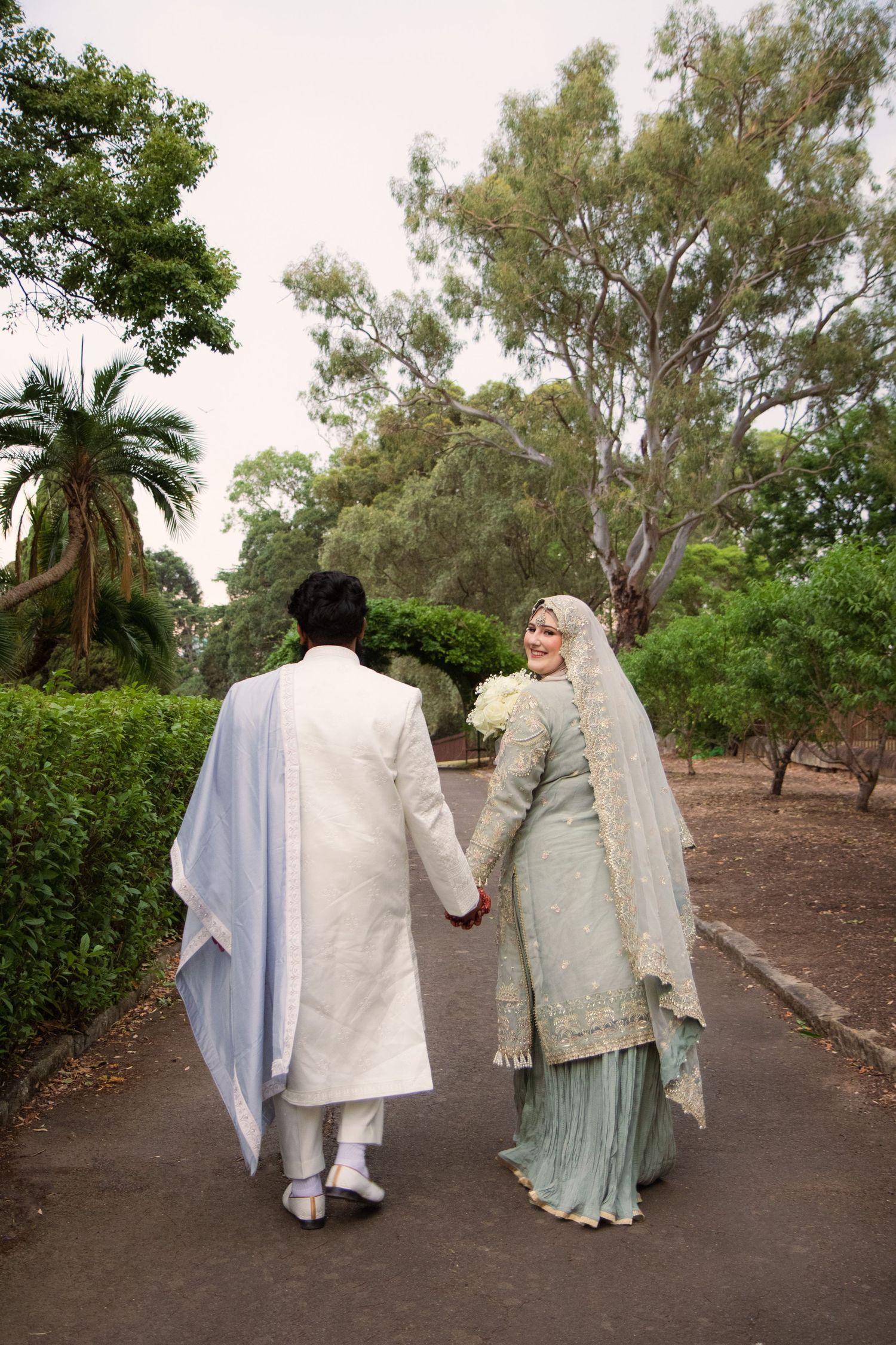 A couple in traditional wedding attire walks down a tree-lined path holding hands in four consecutive photos.