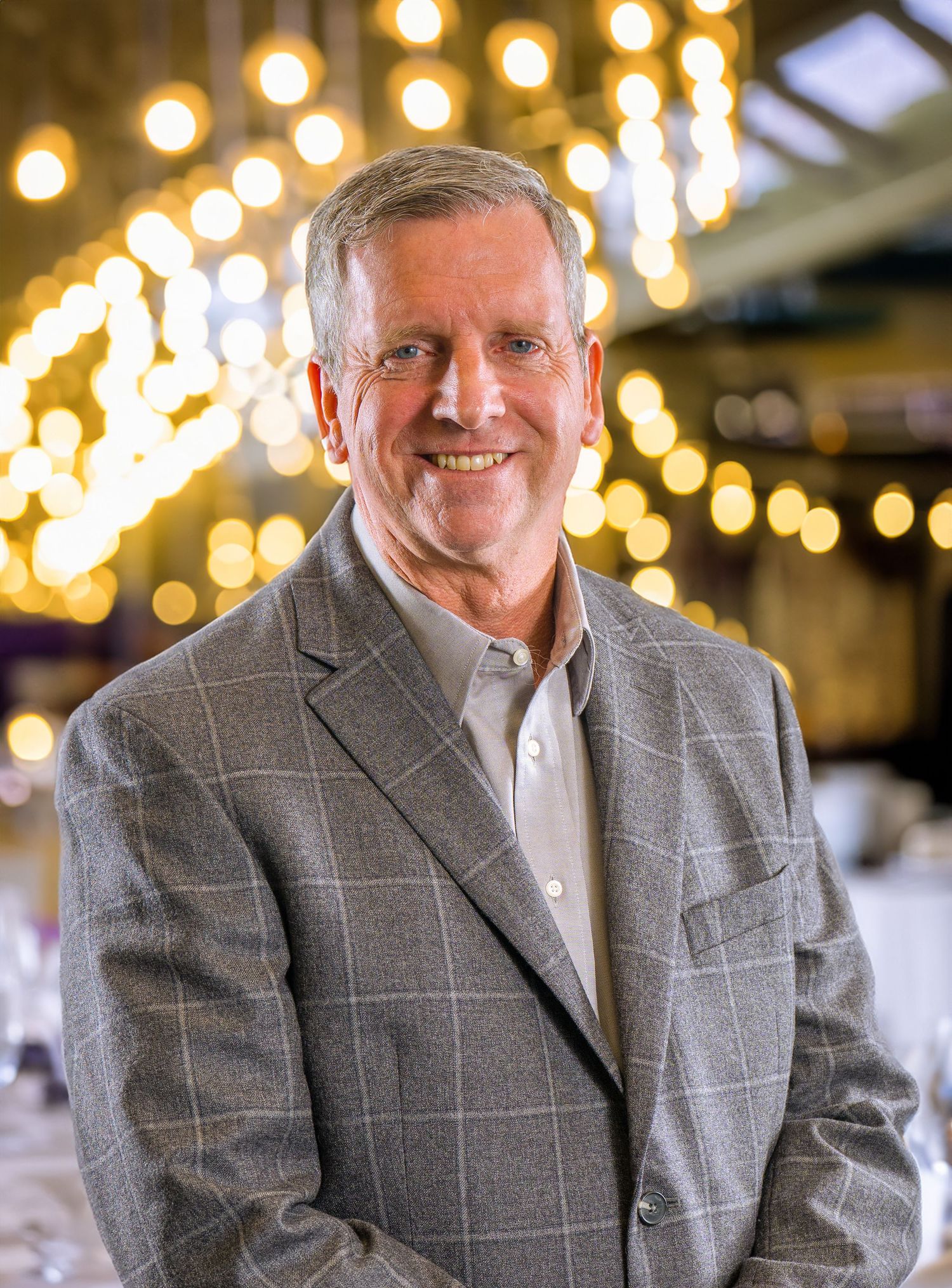Professional business portrait of a person in a gray windowpane blazer smiling warmly against blurred lights background.