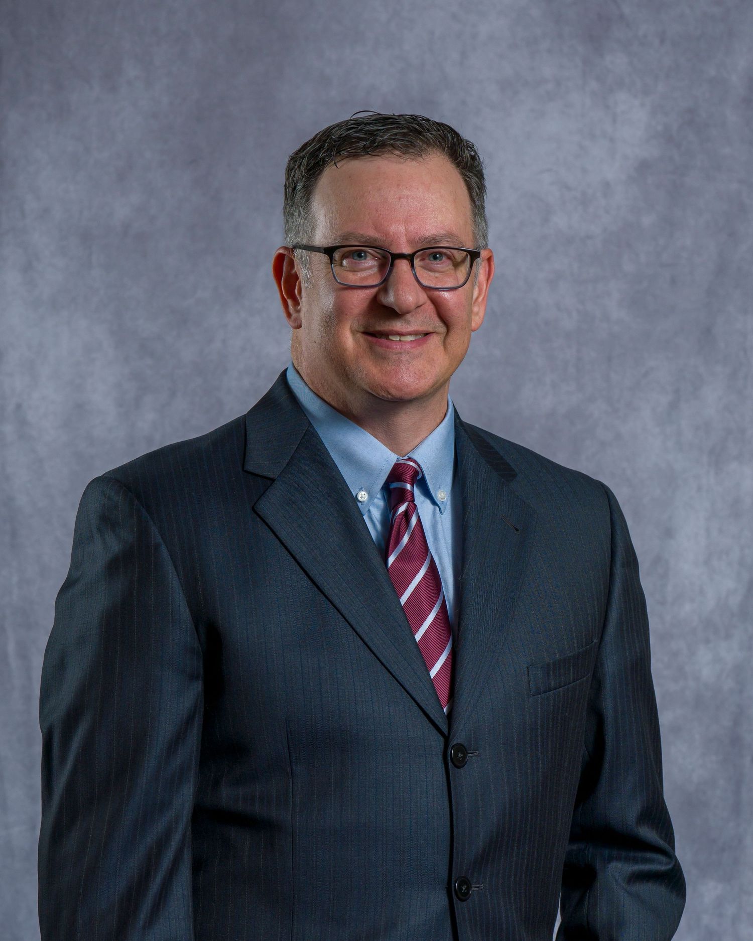 Professional corporate headshot of someone in a dark suit and striped red tie against a gray background.