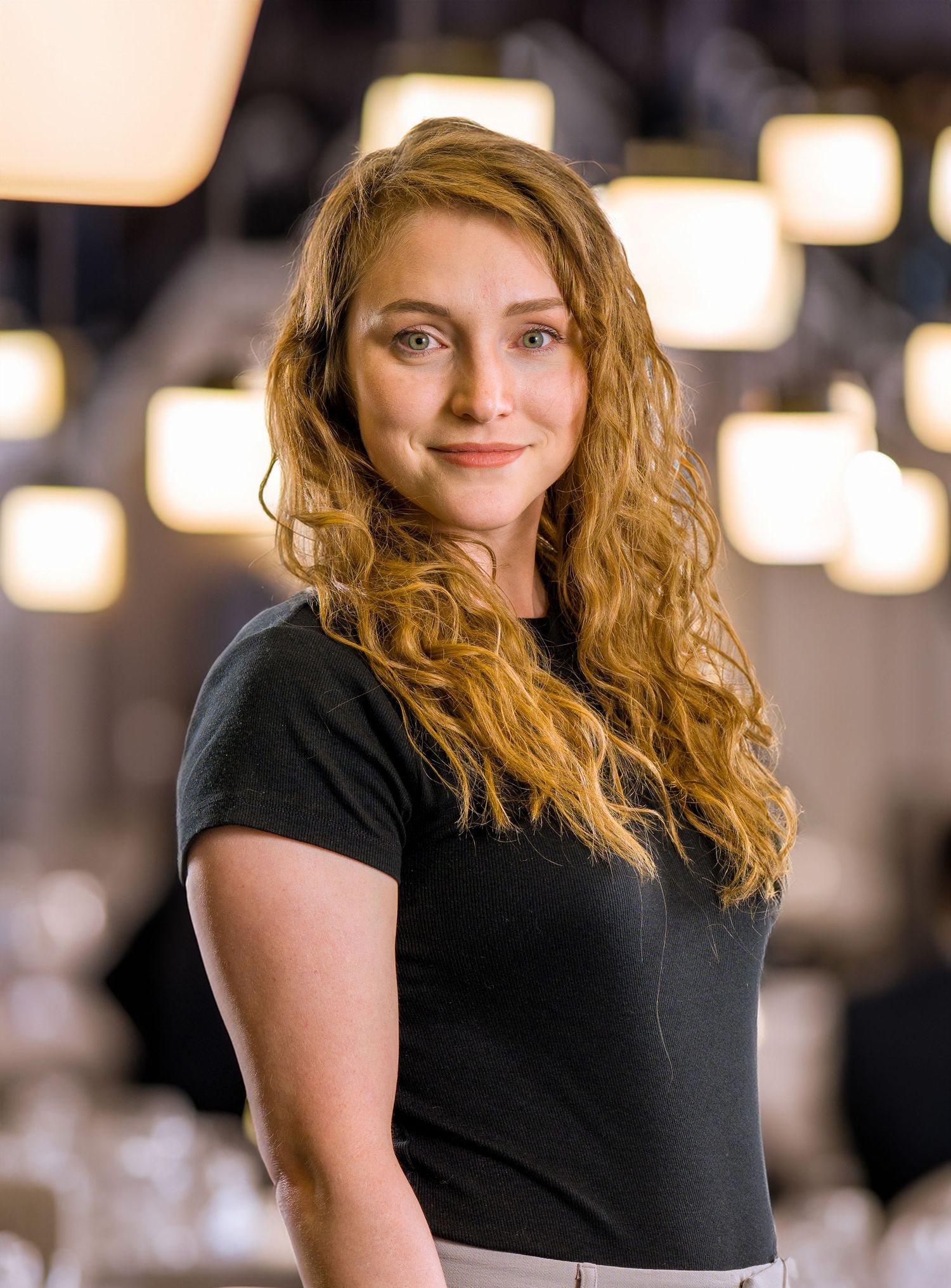 Person with long wavy blonde hair wearing a black t-shirt poses in front of glowing lights in an indoor setting.