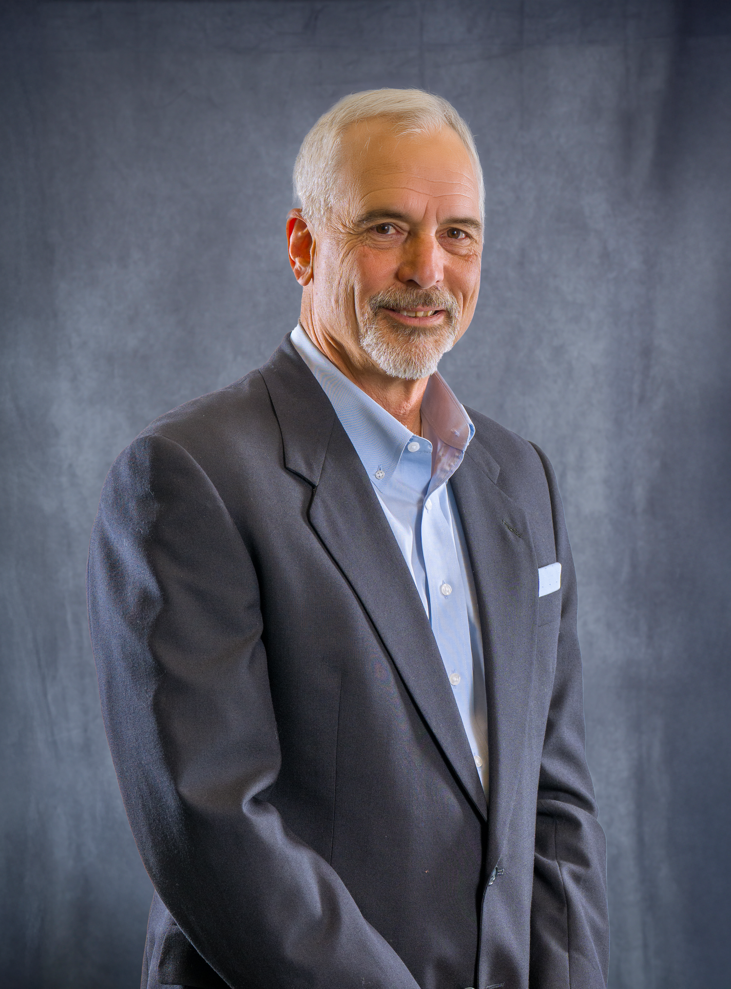 Professional headshot portrait of a smiling senior executive in a dark suit and light blue shirt against a gray background.