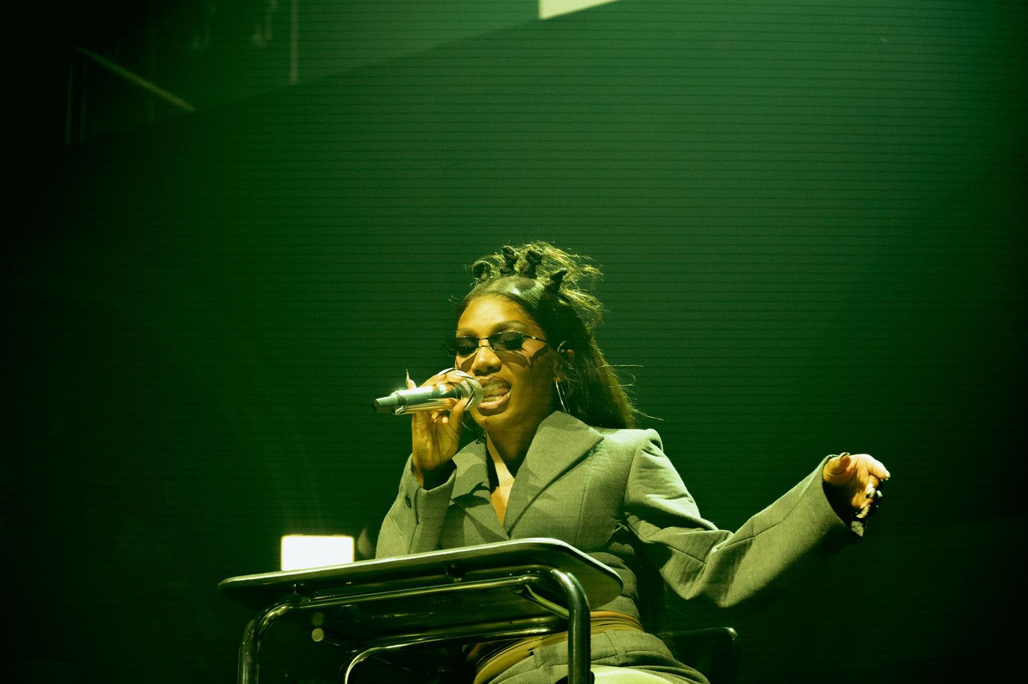 DOECHII in a gray blazer and glasses poses confidently under dramatic stage lighting.
