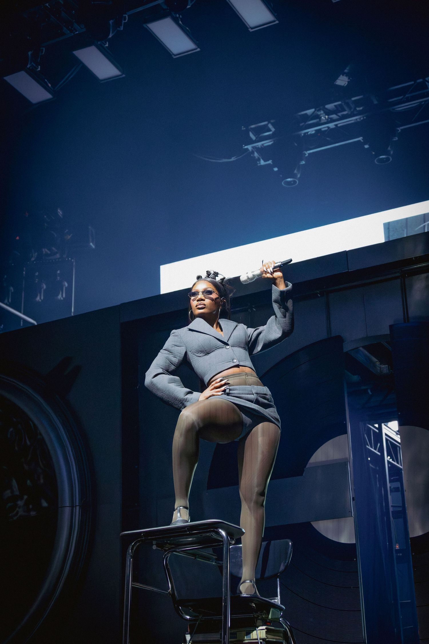 DOECHII in a gray blazer and glasses poses confidently under dramatic stage lighting.