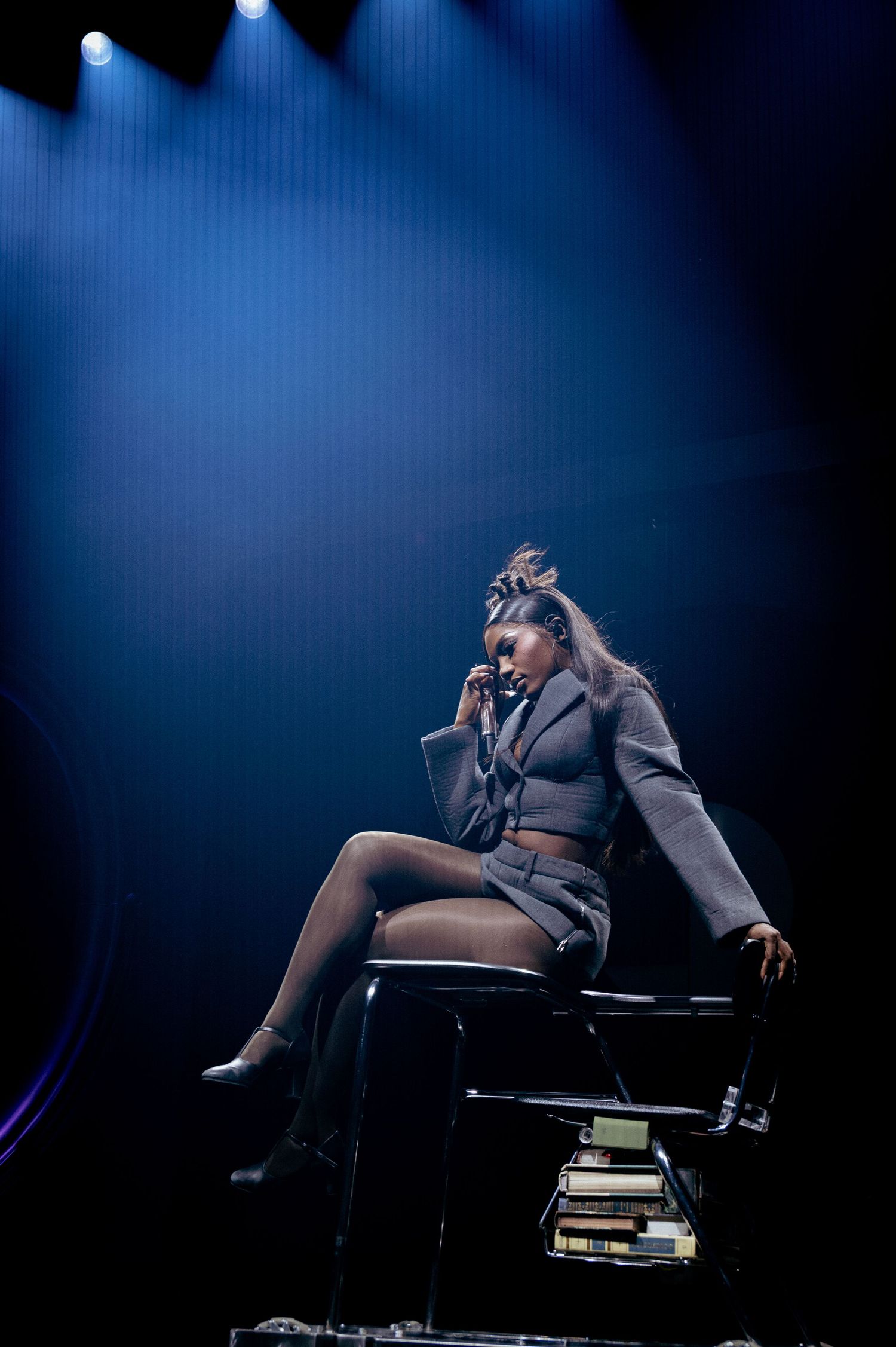 DOECHII in a gray blazer and glasses poses confidently under dramatic stage lighting.
