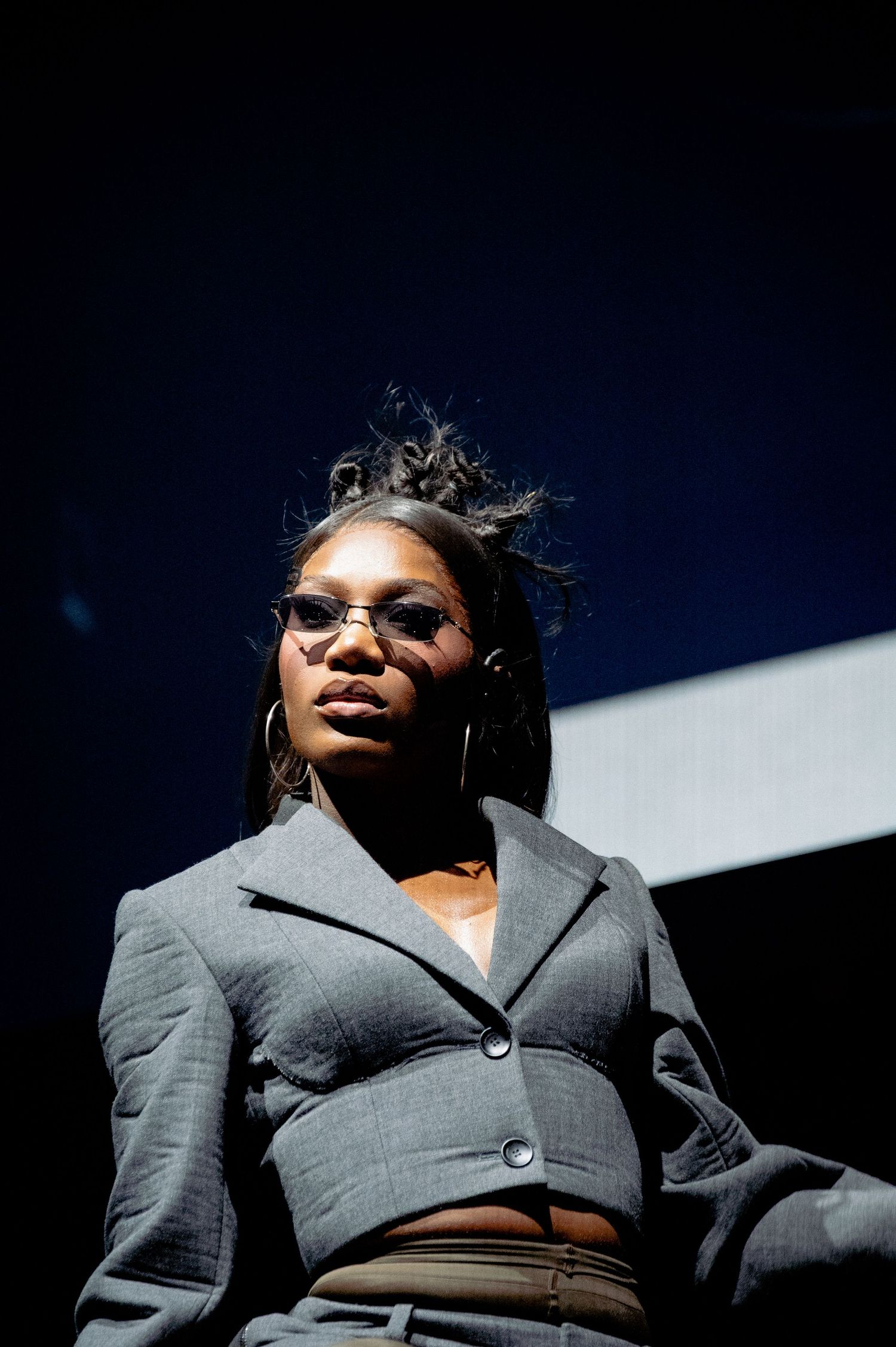 DOECHII in a gray blazer and glasses poses confidently under dramatic stage lighting.