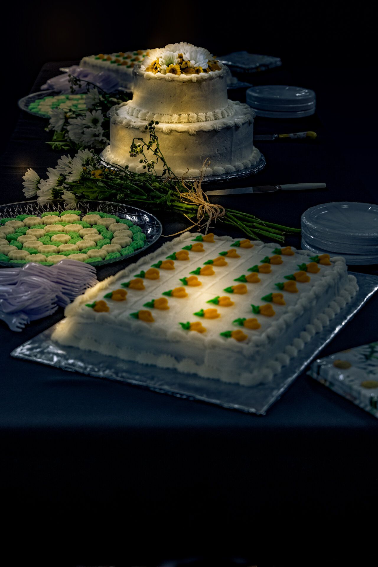 Elegant wedding dessert display with a tiered white cake and decorated sheet cake under dramatic lighting at night.