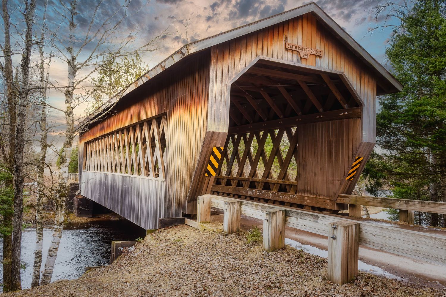 A historic wooden covered bridge spans across a river with dramatic clouds and birch trees in the background.