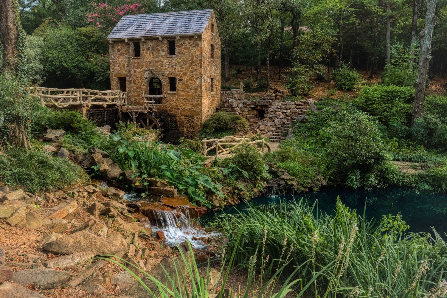 Historic stone mill building with wooden waterwheel perched beside cascading waterfall in lush forest landscape.