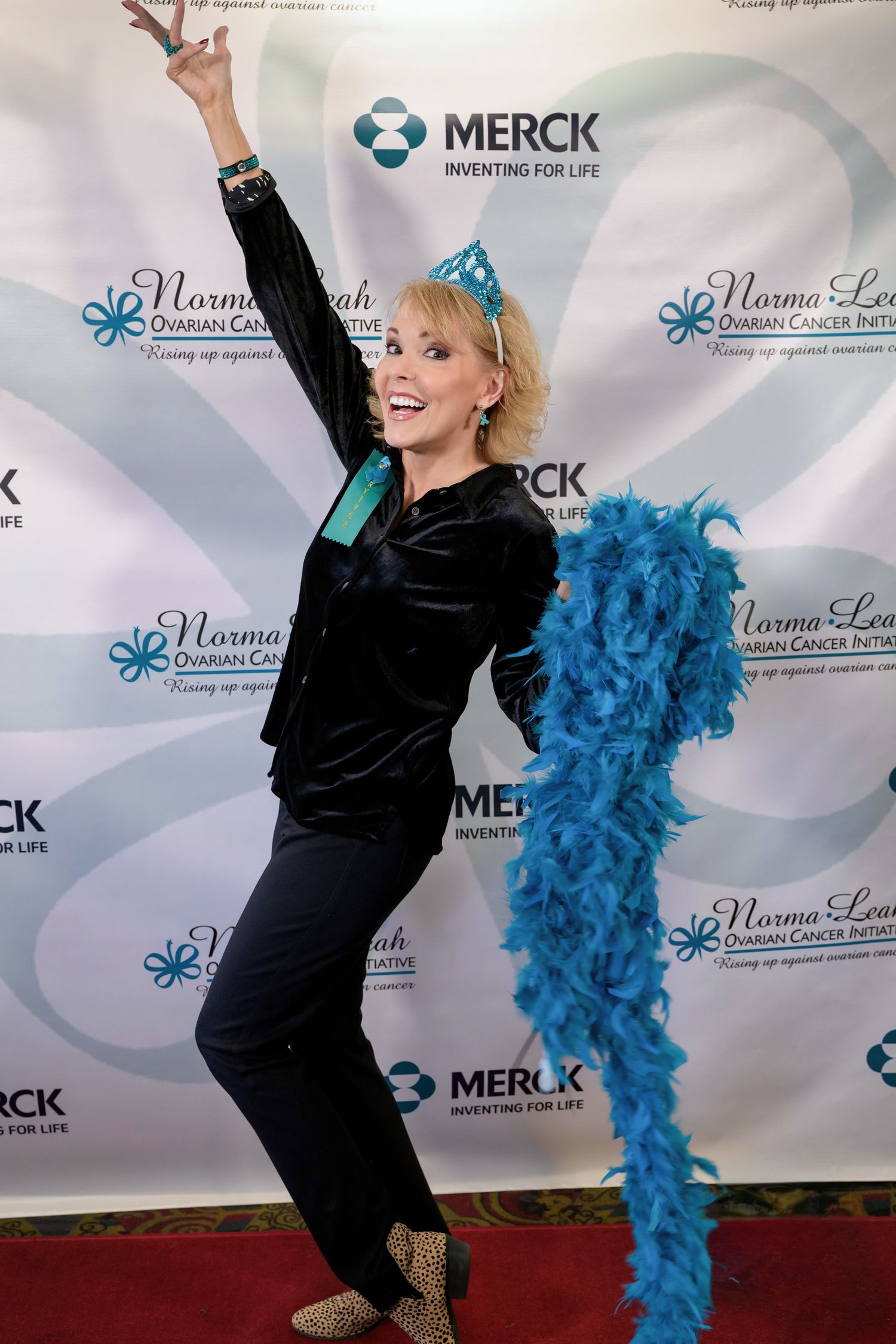 Performer in black outfit strikes energetic pose against Merck sponsor backdrop with blue feather boa at event.